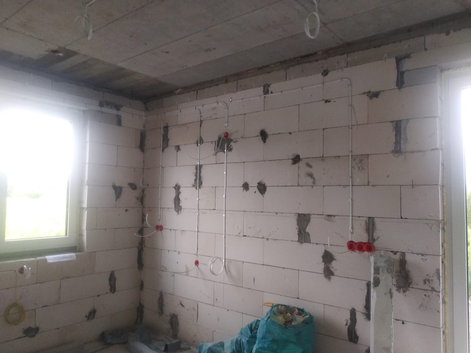 Wnętrze w stanie surowym: białe ściany z betonu komórkowego, widoczne przewody elektryczne w białych rurkach, czerwone puszki instalacyjne. W tle okno z widokiem na zieleń.
