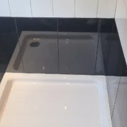 Biały brodzik kabiny prysznicowej z czarną i białą glazurą na ścianach i podłodze, widoczne fugi i odpływ.