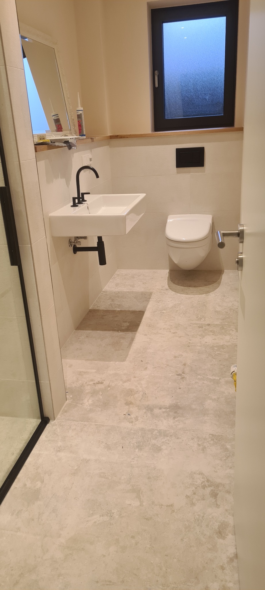 Nowoczesna łazienka z podwieszaną umywalką z czarną armaturą, toaletą w zabudowie, lustrem i oknem w czarnej ramie, wykończona płytkami w odcieniach beżu i szarości.