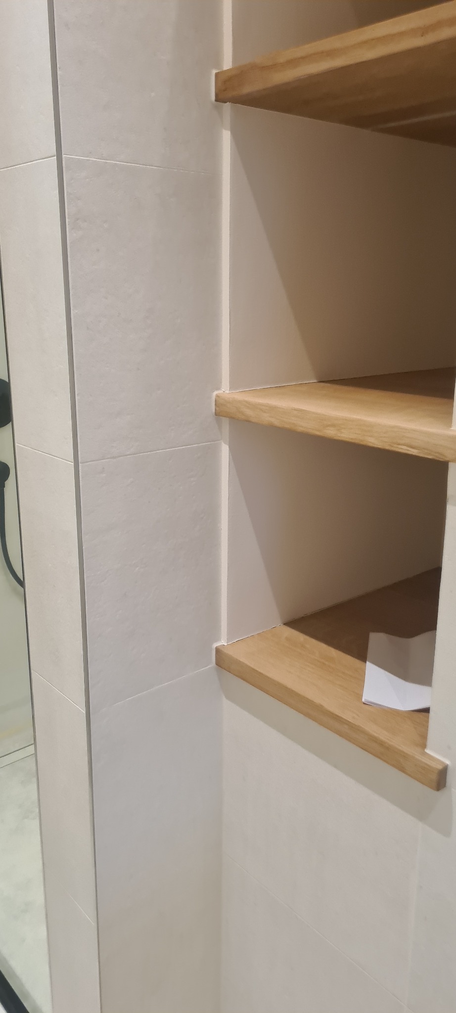 Wnęka w ścianie wyłożona białymi płytkami, z trzema drewnianymi półkami. Na dolnej półce leży złożona biała kartka papieru.