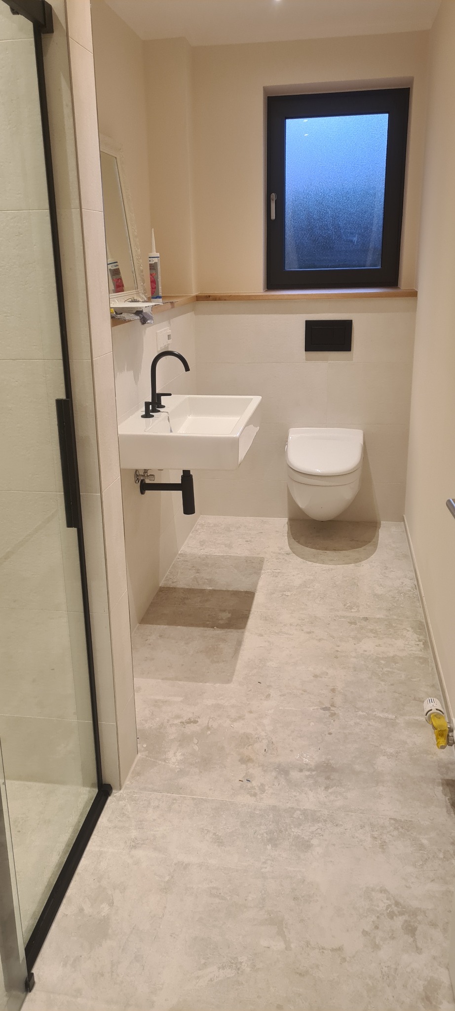 Nowoczesna łazienka z prostokątną umywalką nablatową i czarną armaturą, wisząca toaleta, szare płytki podłogowe, okno z czarną ramą i częściowo widoczna kabina prysznicowa.