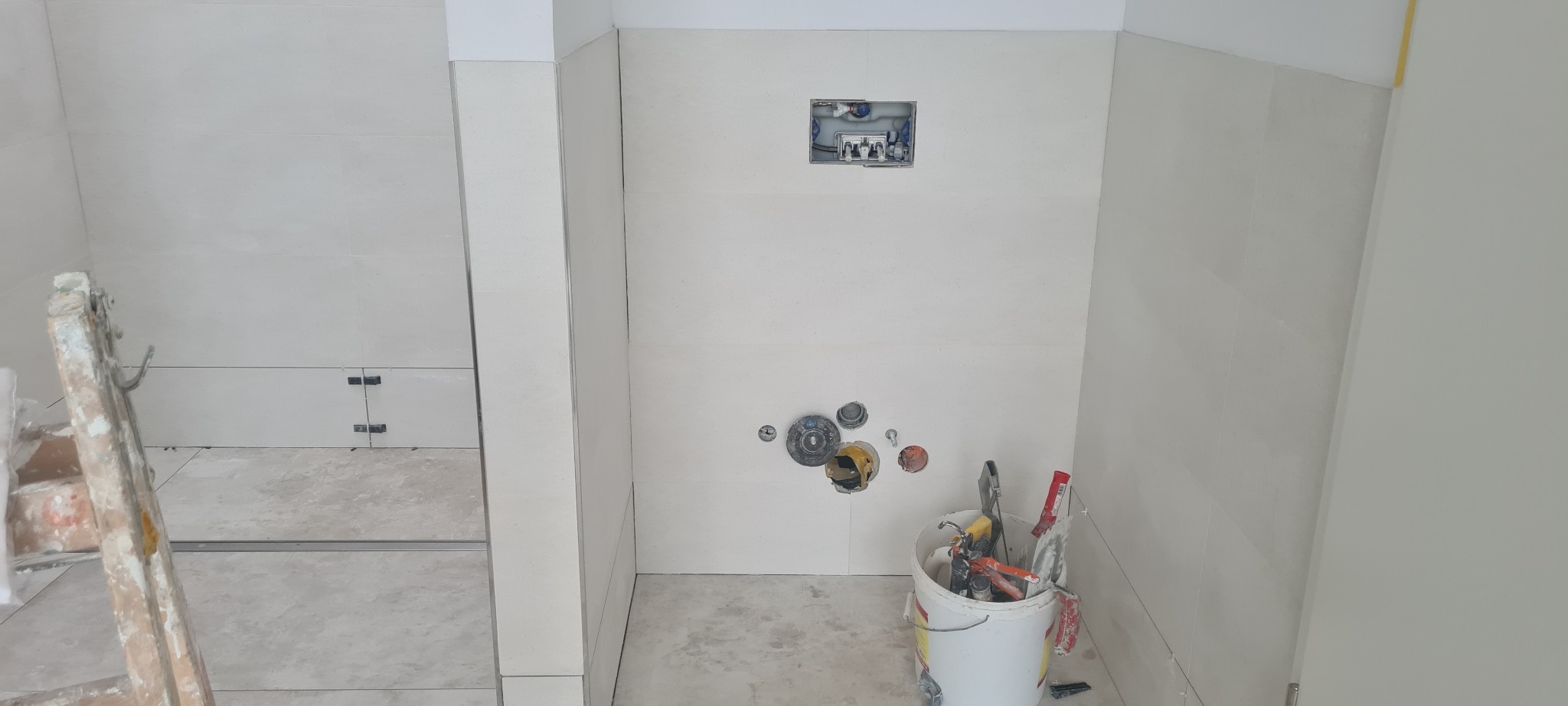 Świeżo wykafelkowana łazienka w trakcie prac wykończeniowych; widoczne otwory na armaturę, białe płytki, wiadro z narzędziami i drabina malarska.