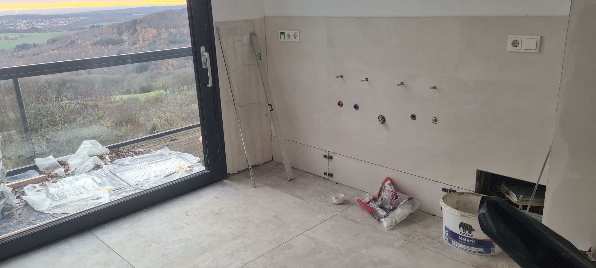 Wnętrze w trakcie wykańczania: ułożone płytki na ścianie z przygotowanymi punktami hydraulicznymi, poziomica oparta o ścianę, przy oknie zabezpieczone płytki folią i resztkami liści, widok...