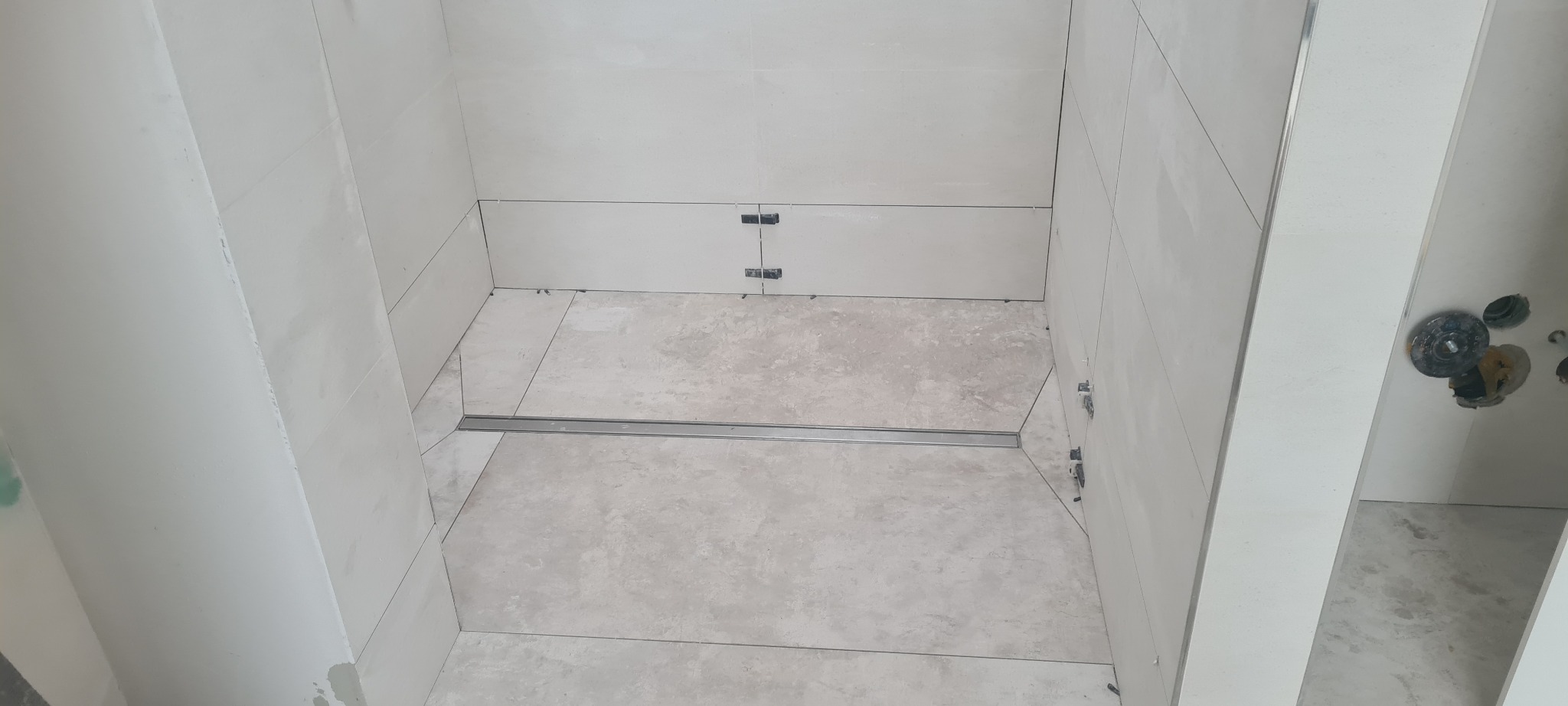 Łazienka w trakcie wykańczania: jasne płytki ścienne częściowo ułożone, widoczny odpływ liniowy w podłodze, elementy montażowe na ścianach, surowy stan instalacji oświetleniowej.