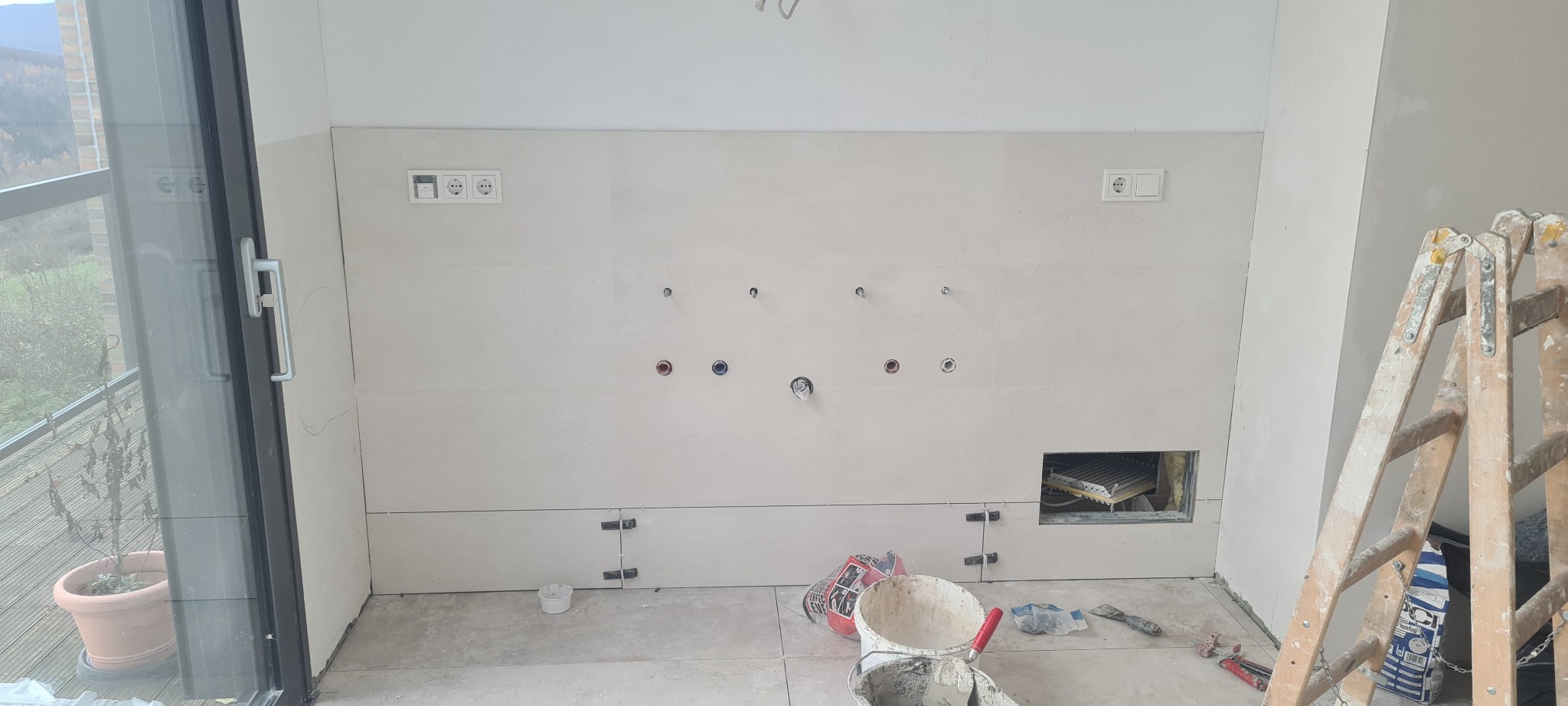 Ściana w trakcie układania płytek z widocznymi puszkami elektrycznymi, rurami, poziomymi listwami dystansowymi, drabina malarska i narzędzia glazurnicze na podłodze, balkon z widokiem na zalesione...