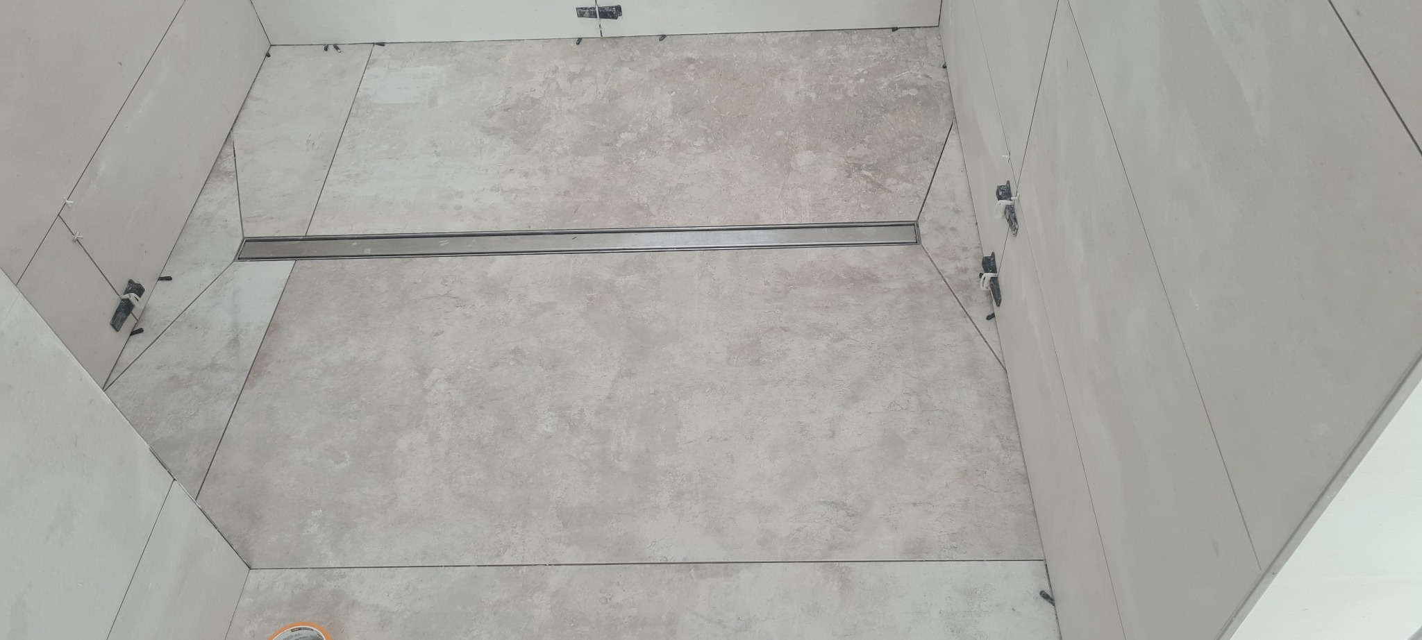 Ułożone płytki podłogowe w kabinie prysznicowej z odpływem liniowym, widoczne krzyżyki dystansowe i poziomica laserowa na ścianie, jasne płytki imitujące kamień.