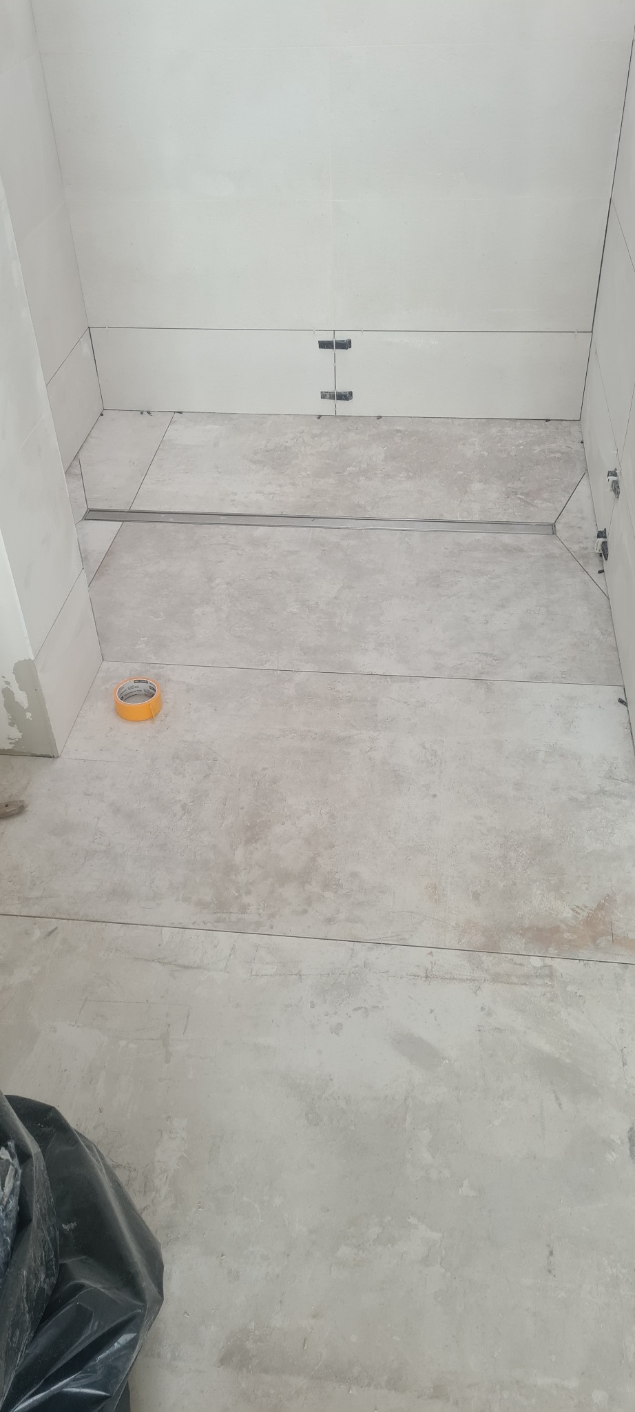 Wnętrze łazienki w trakcie układania płytek podłogowych i ściennych, widoczny odpływ liniowy, taśma malarska i system poziomowania płytek.