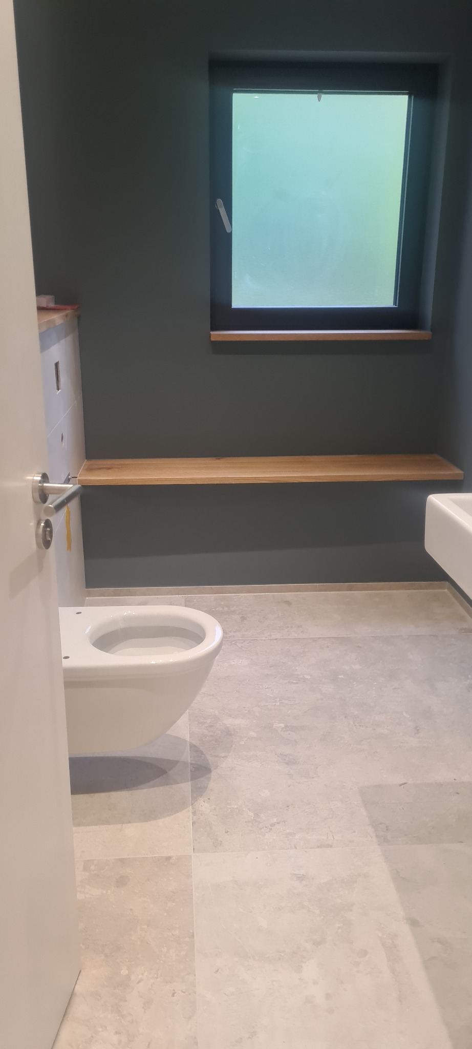Nowoczesna łazienka z podwieszaną toaletą, drewnianą półką pod oknem i szarymi płytkami podłogowymi, widok zza uchylonych drzwi.