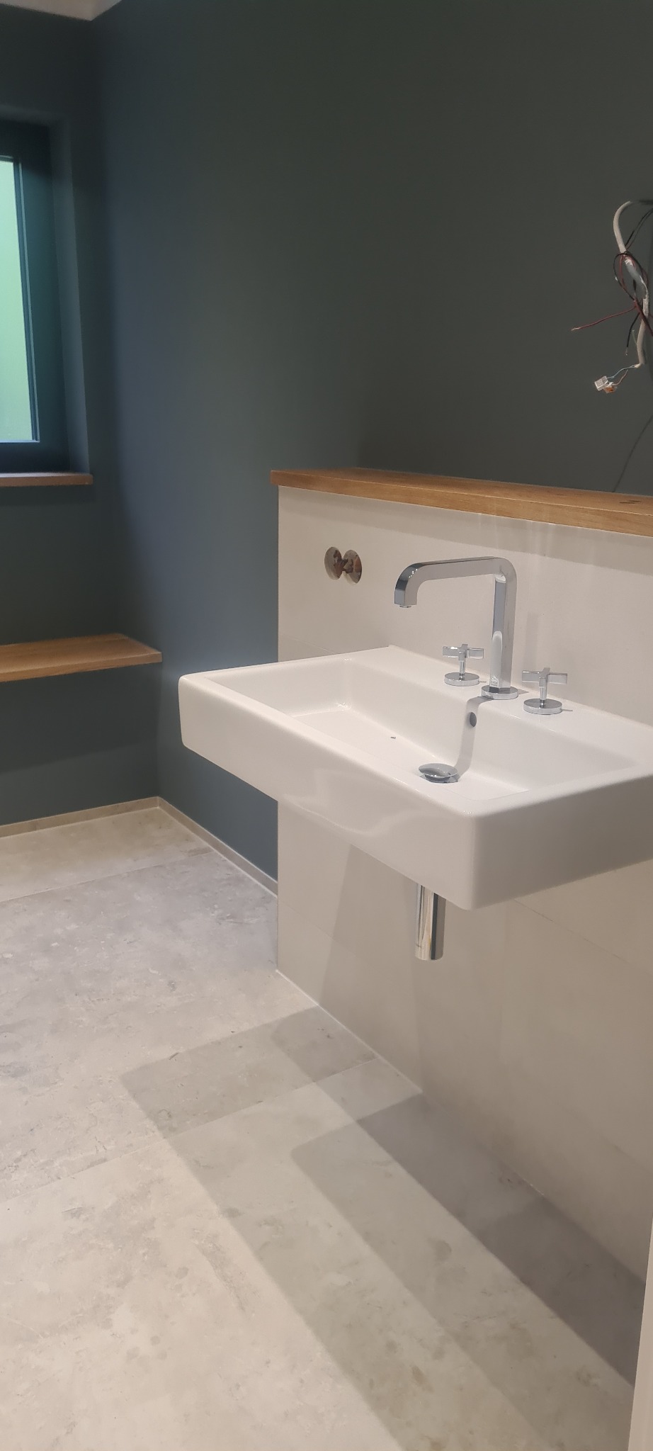 Nowoczesna, kwadratowa umywalka z chromowaną armaturą krzyżową na tle ściany w kolorze ciemnego turkusu, z drewnianym blatem i półkami w łazience wykończonej płytkami imitującymi kamień.