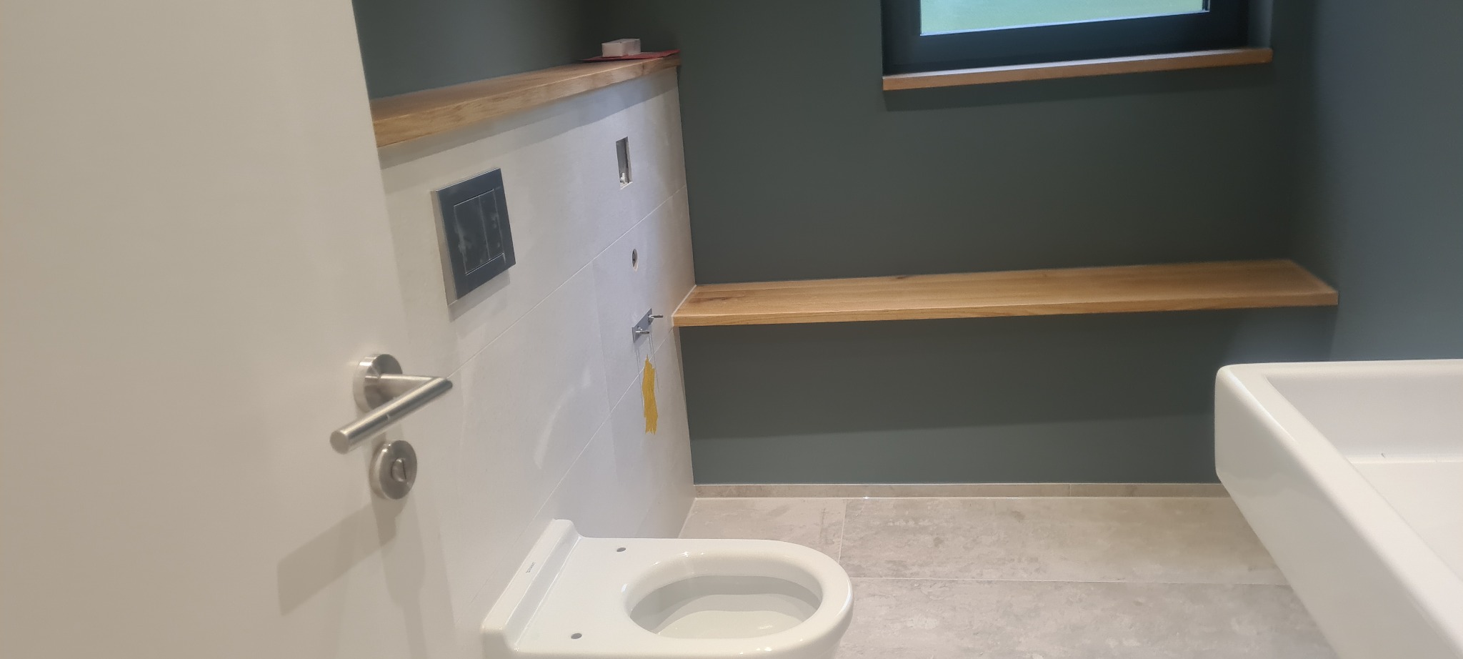 Nowoczesna łazienka w trakcie wykańczania: podwieszana toaleta, białe płytki z drewnianymi półkami, szara ściana i podłoga.