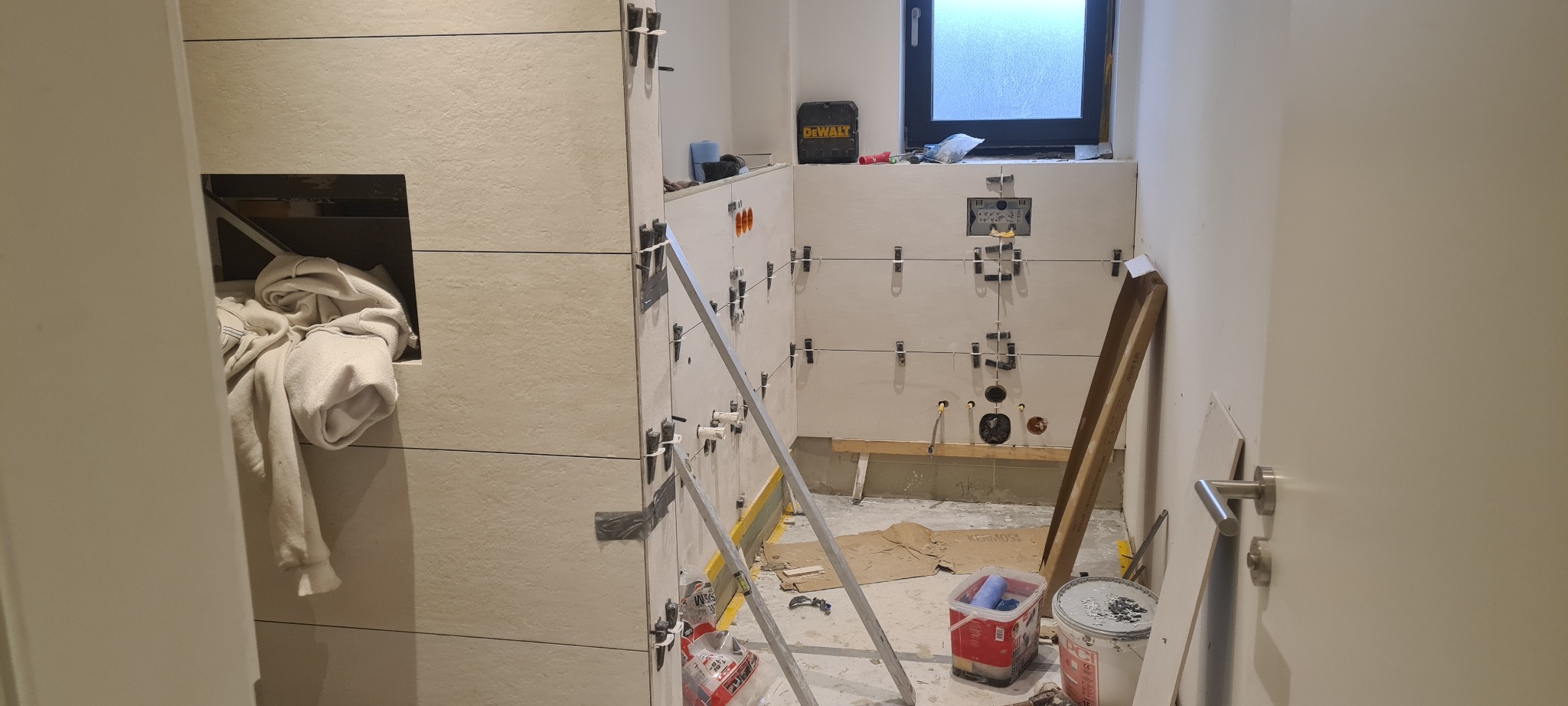 Łazienka w trakcie układania płytek ściennych, widoczne krzyżyki dystansowe, drabina, narzędzia, otwór na półkę z ręcznikiem i okno.