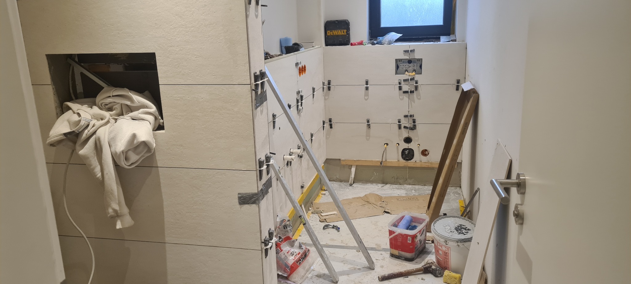 Łazienka w trakcie układania płytek; widoczne poziomice, plastikowe krzyżyki dystansowe, narzędzia glazurnicze i ubranie w niszy ściennej.