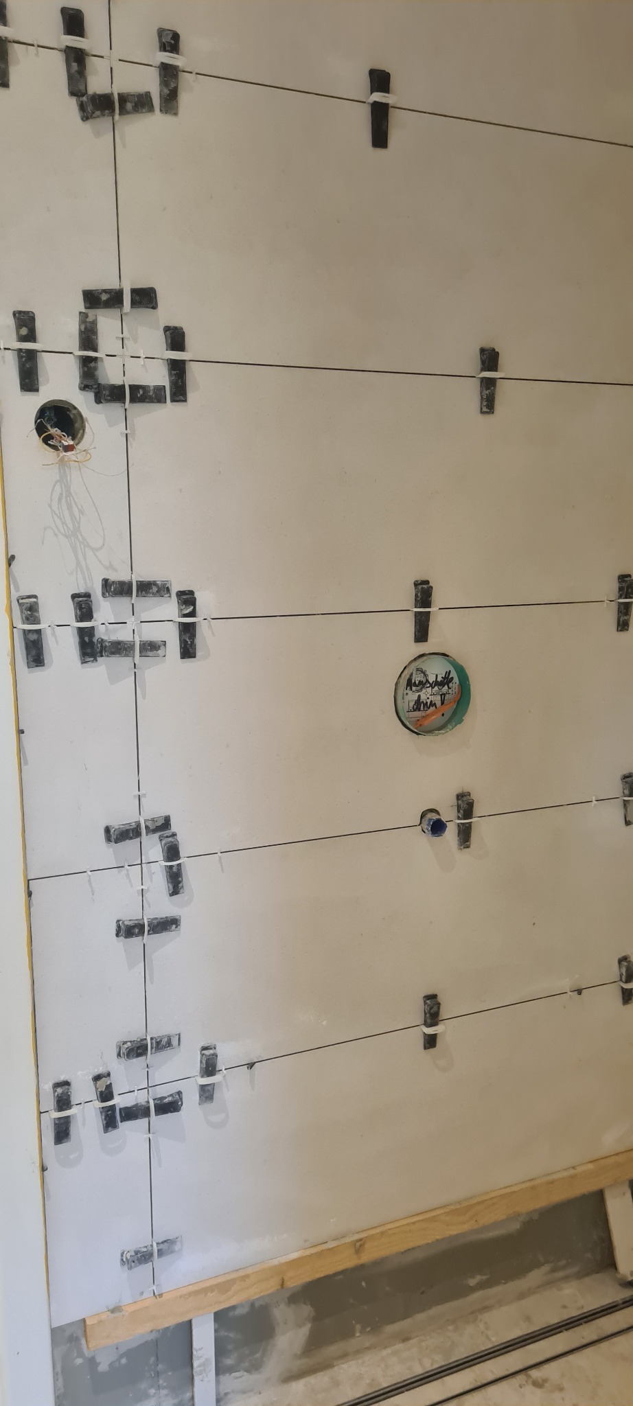 Ściana w trakcie układania płytek, widoczne krzyżyki dystansowe, sznurki poziomujące, otwory na gniazdka elektryczne i rury.
