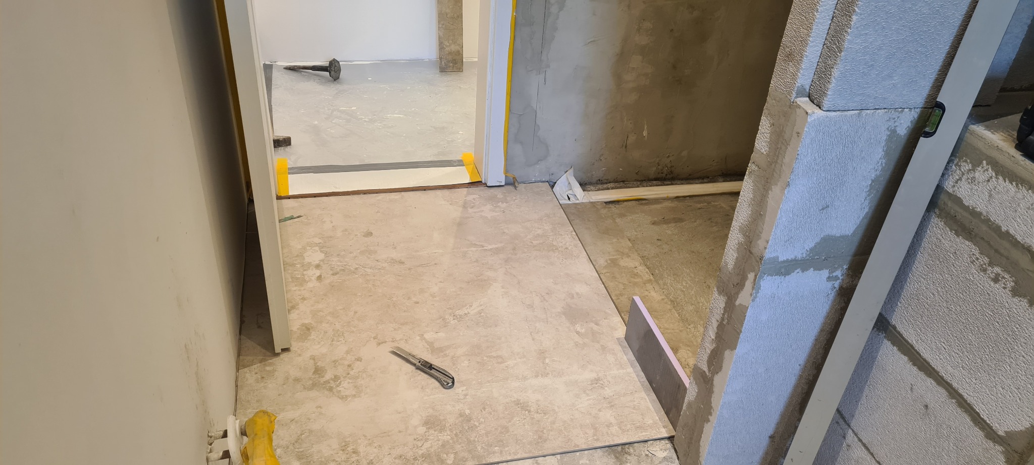 Układanie płytek podłogowych w trakcie prac wykończeniowych, widoczny nóż do cięcia, poziomica przy ścianie z bloczków, zabezpieczenia folią malarską w przejściu do innego pomieszczenia.