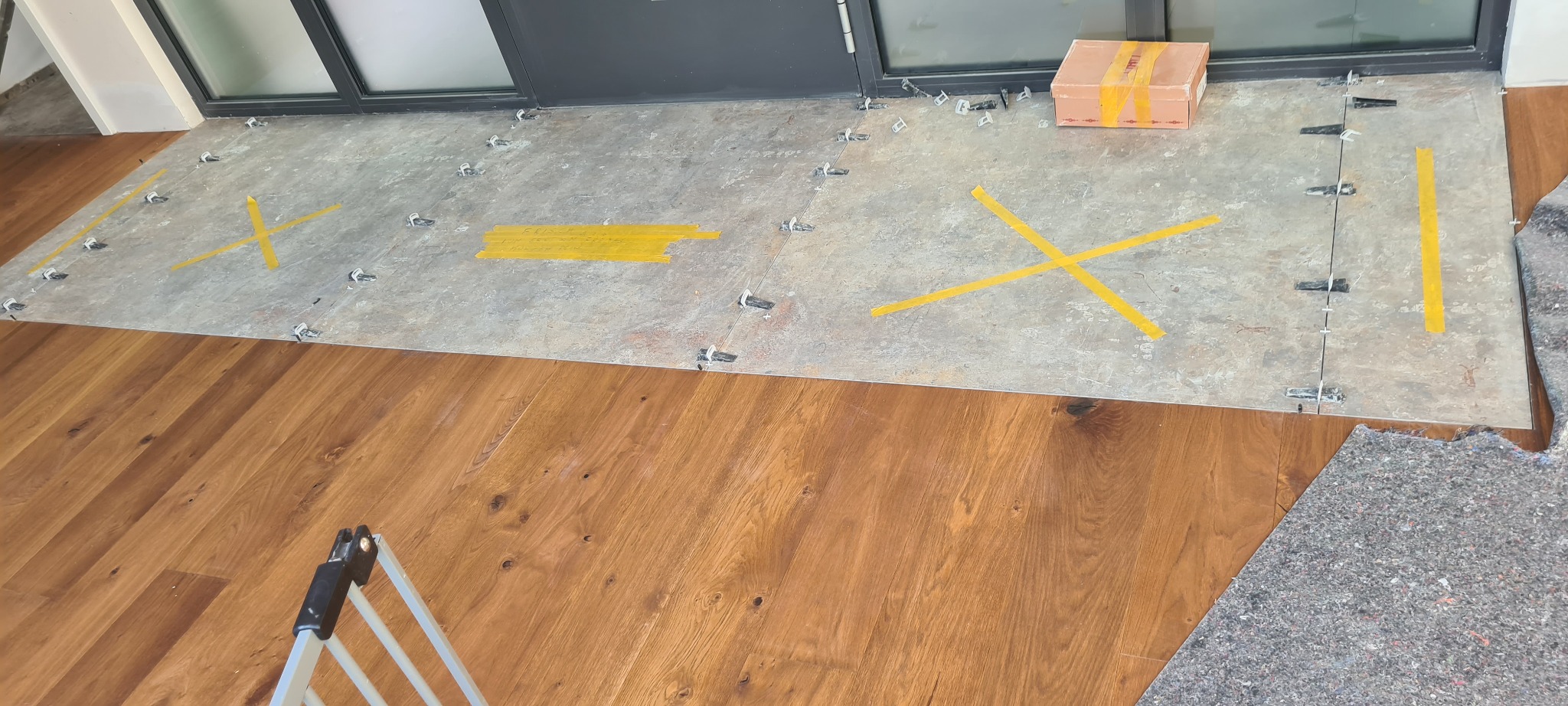 Przygotowanie do ułożenia płytek podłogowych: szare płytki z żółtą taśmą wyznaczającą obszary, plastikowe krzyżyki dystansowe, kartonowe pudełko, fragment drewnianej podłogi i drabinka w kadrze.