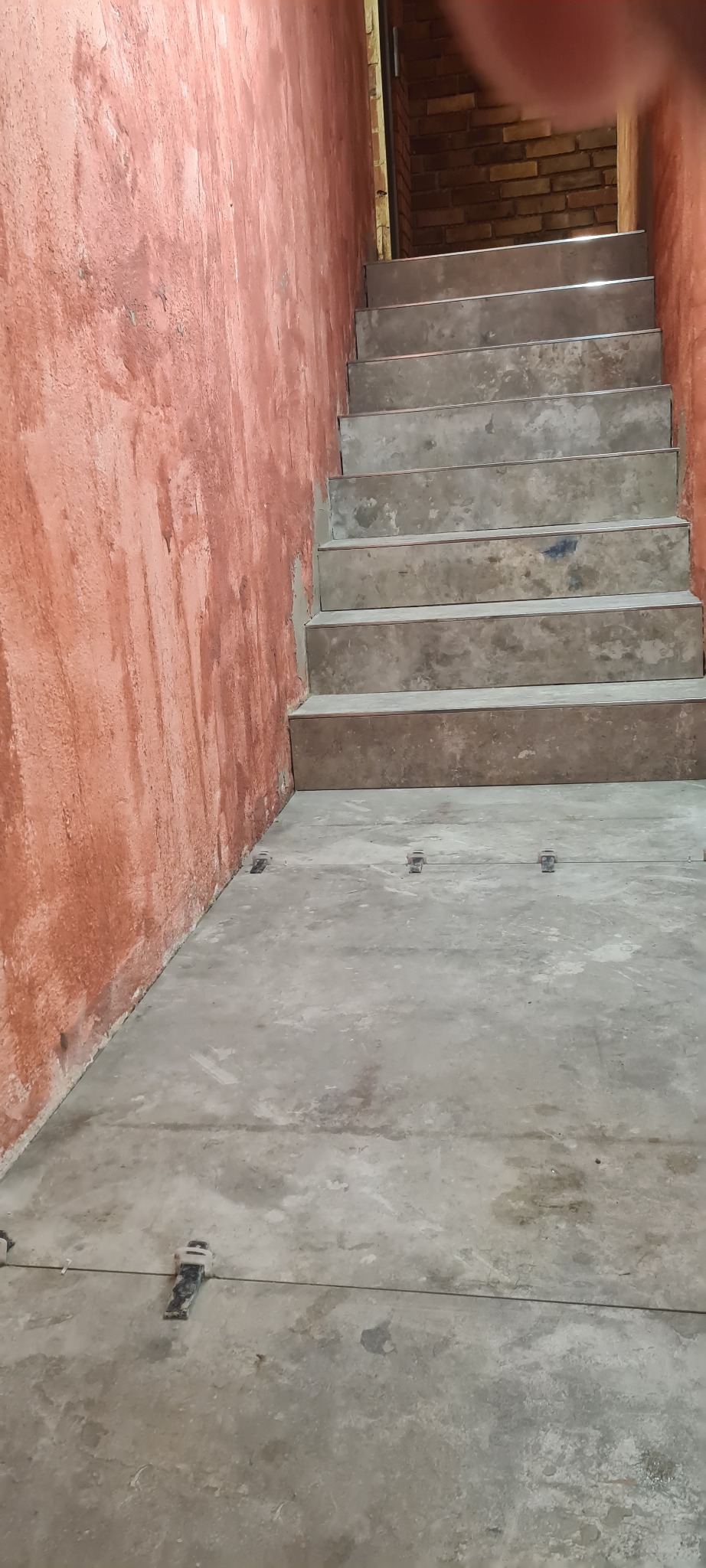 Surowe betonowe schody prowadzące do piwnicy lub innego pomieszczenia, pomalowana na czerwono ściana z widoczną fakturą, widoczne ślady po niedokładnym malowaniu, na podłodze widoczne elementy...