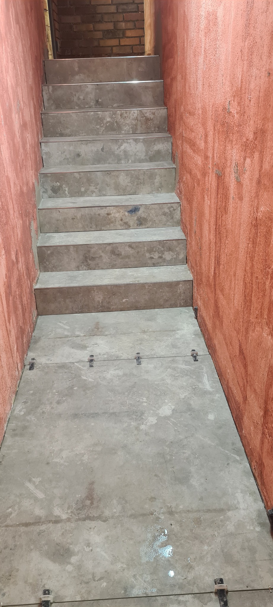 Surowe betonowe schody w klatce schodowej z pomarańczowymi ścianami, widoczne metalowe elementy mocujące na podłodze.