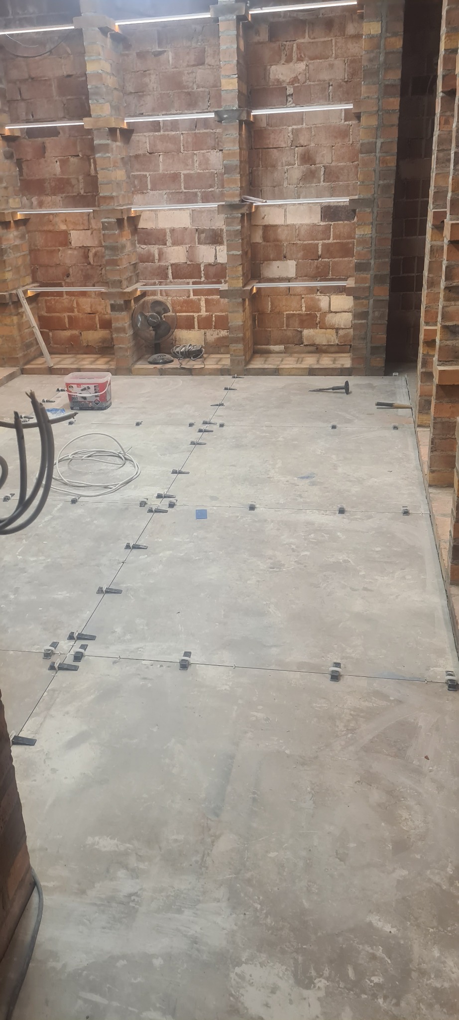 Przygotowanie podłoża pod płytki z użyciem krzyżyków dystansowych, widoczny mur z cegły, wentylator i narzędzia.