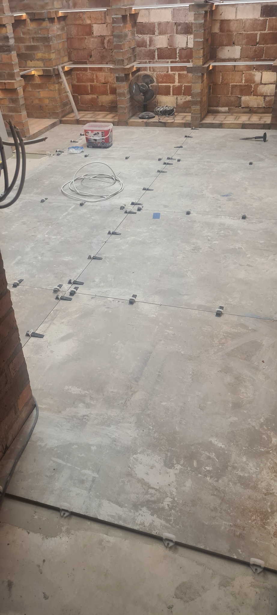 Przygotowanie podłoża pod płytki: widoczne listwy i klipsy poziomujące na szarej, surowej wylewce, w tle ceglana ściana i wiadro z klejem.