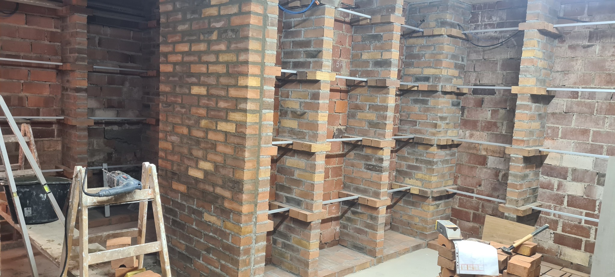 Murowanie z cegły klinkierowej wnęk w ścianie, wsporniki metalowe, drabina, wiadro i wiertarka w tle.