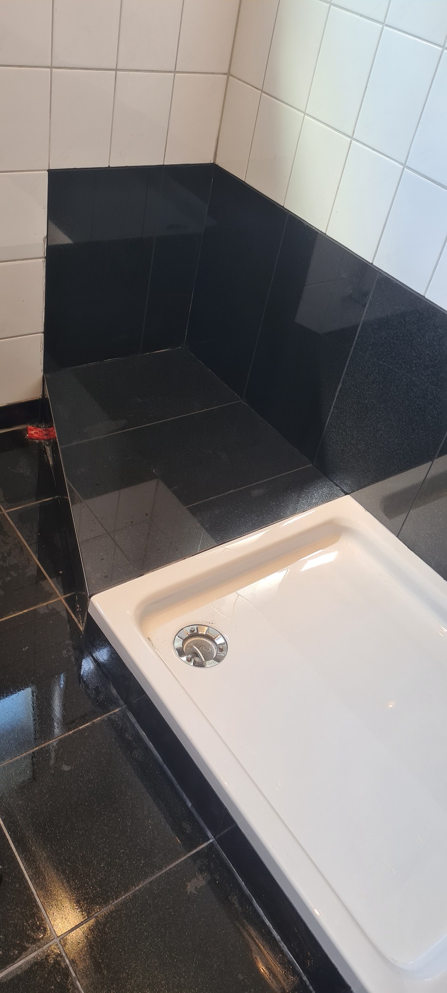 Biały brodzik z metalowym odpływem, otoczony czarnymi i białymi płytkami w łazience, widoczne ślady fugowania.