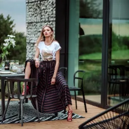 Blondwłosa modelka w szerokiej spódnicy w paski i białej koszulce siedzi na krześle przy stole na drewnianym tarasie, na którym leży czarno-biały dywan, w tle nowoczesny dom z elewacją z kamienia...