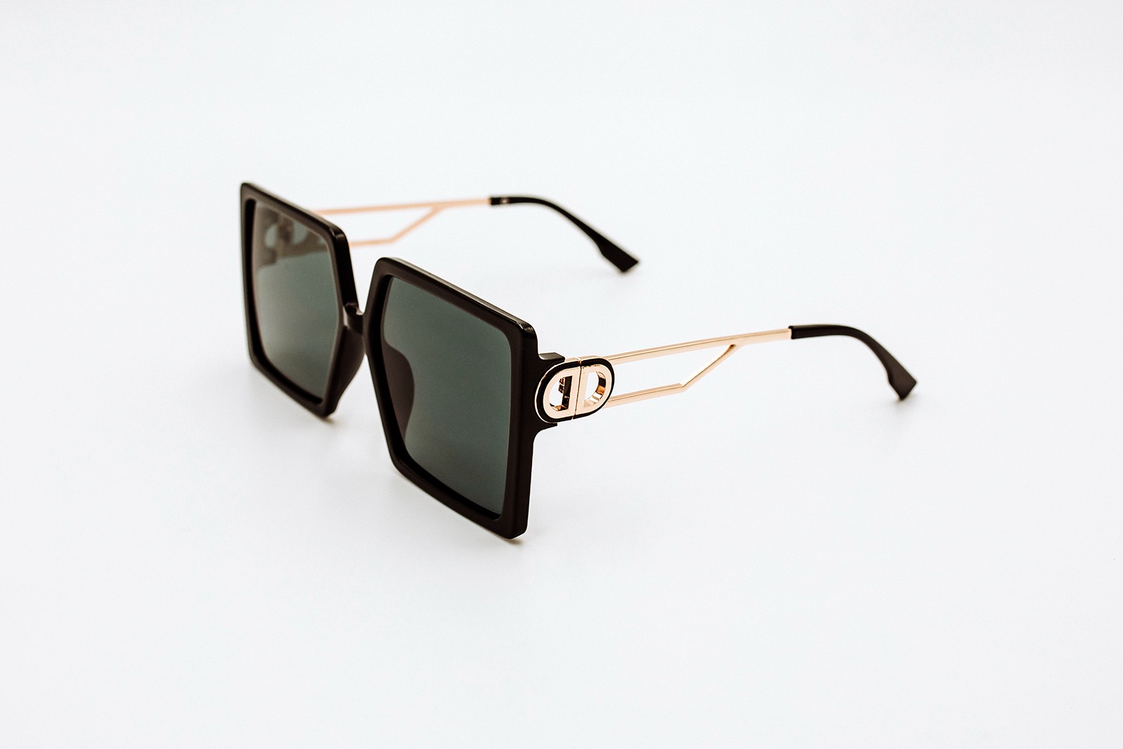Czarne, kwadratowe okulary przeciwsłoneczne z złotymi zausznikami i logo na białym tle.