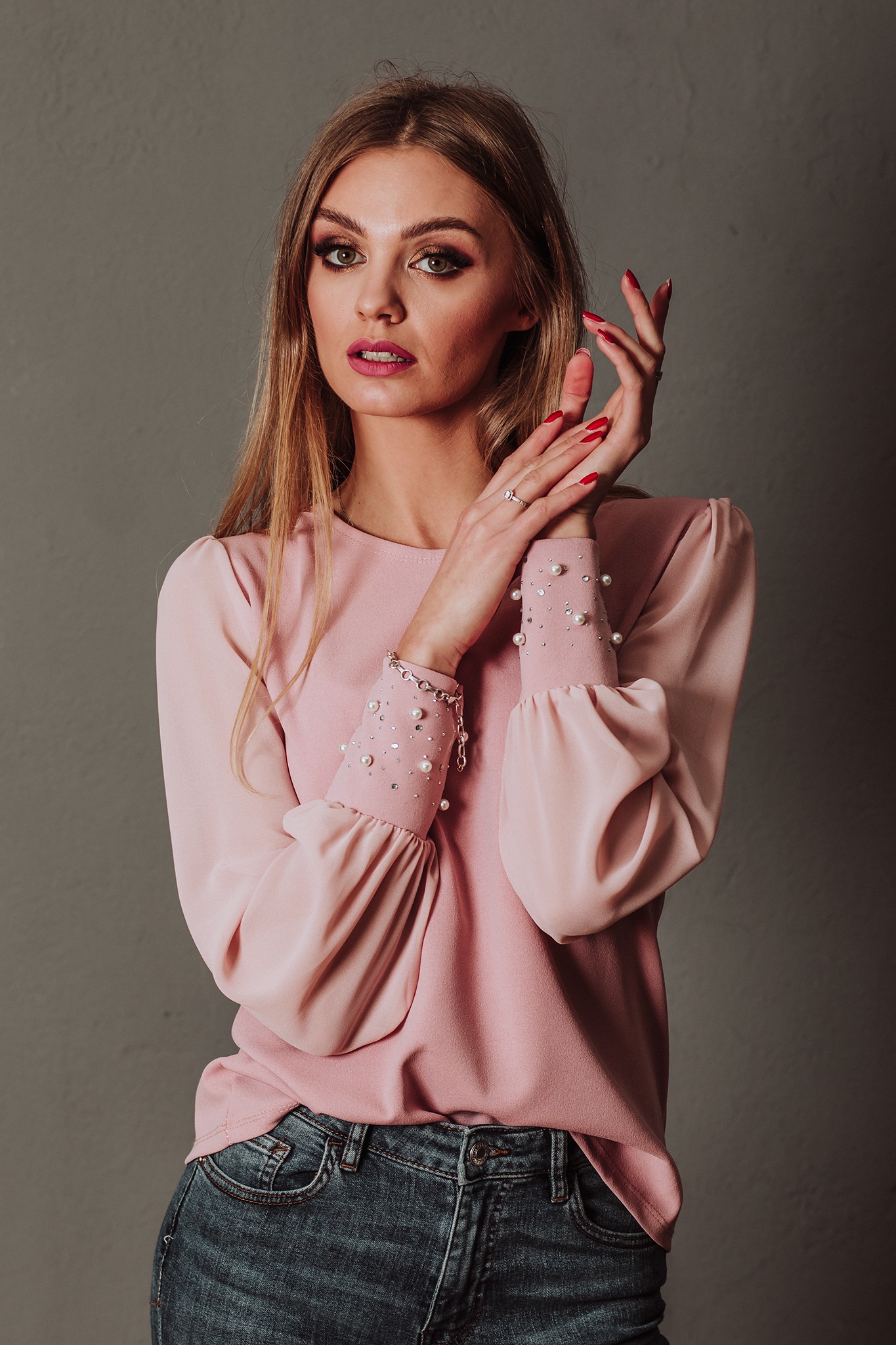 Portret kobiety w studio, ubrana w różową bluzkę z perłami i dżinsy, z rękami uniesionymi w eleganckim geście, na szarym tle.