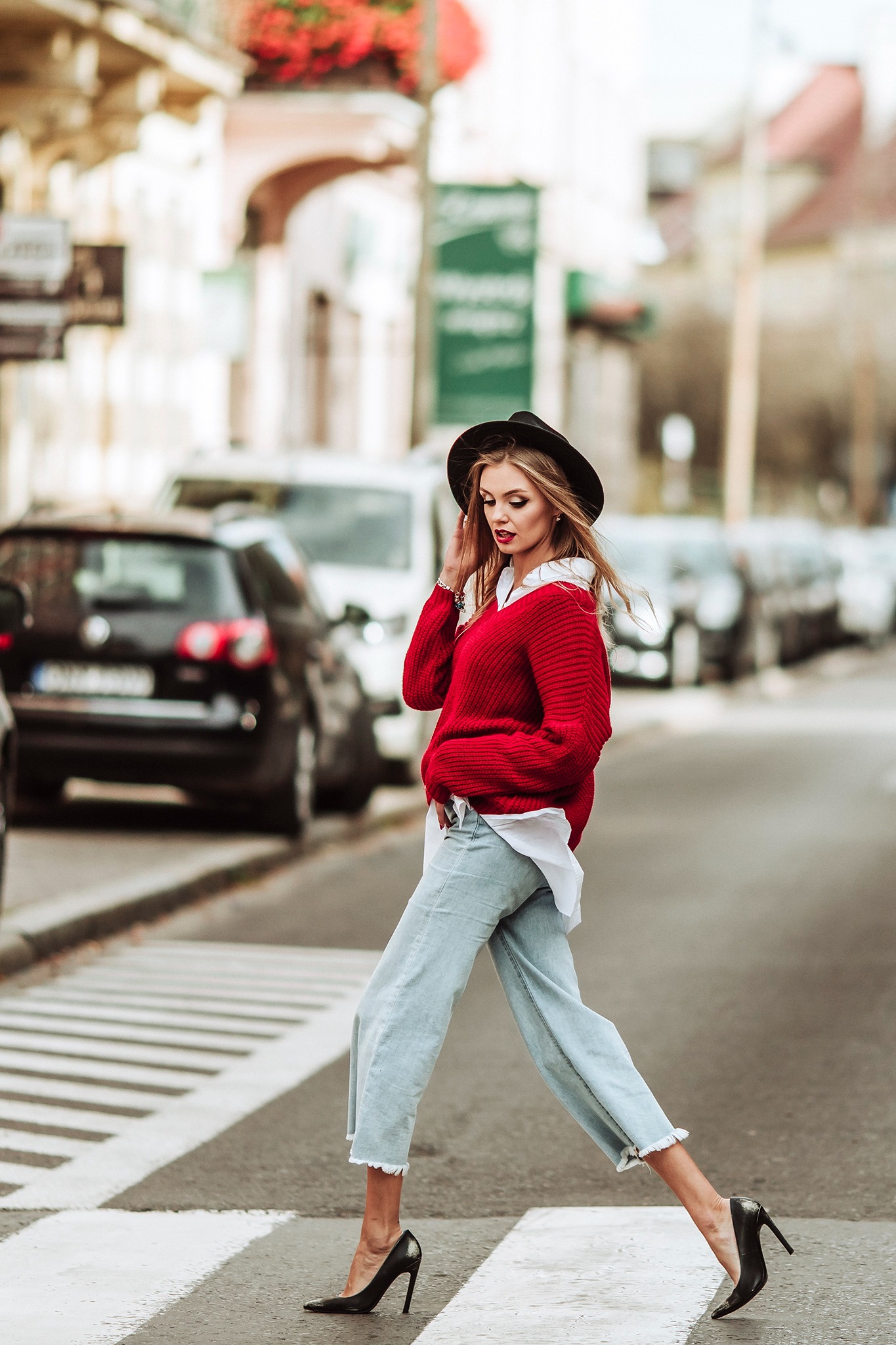 Stylowa blondynka w czerwonym swetrze, białej koszuli, szerokich jeansach i czarnych szpilkach przechodzi przez pasy na tle ulicy z samochodami.