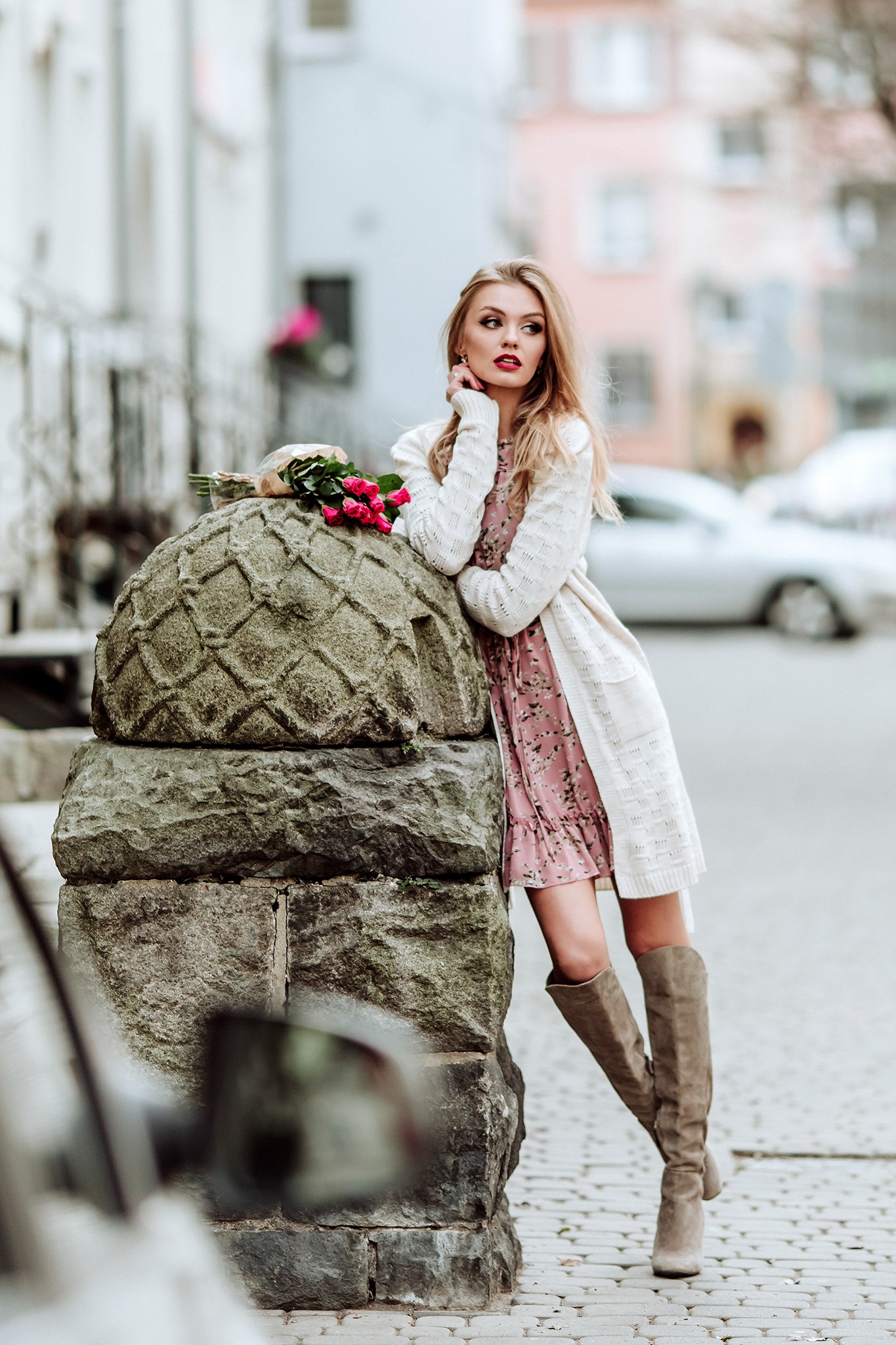 Modelka w kwiecistej sukience i długim swetrze opiera się o kamienny postument z bukietem róż, pozując w miejskiej scenerii.