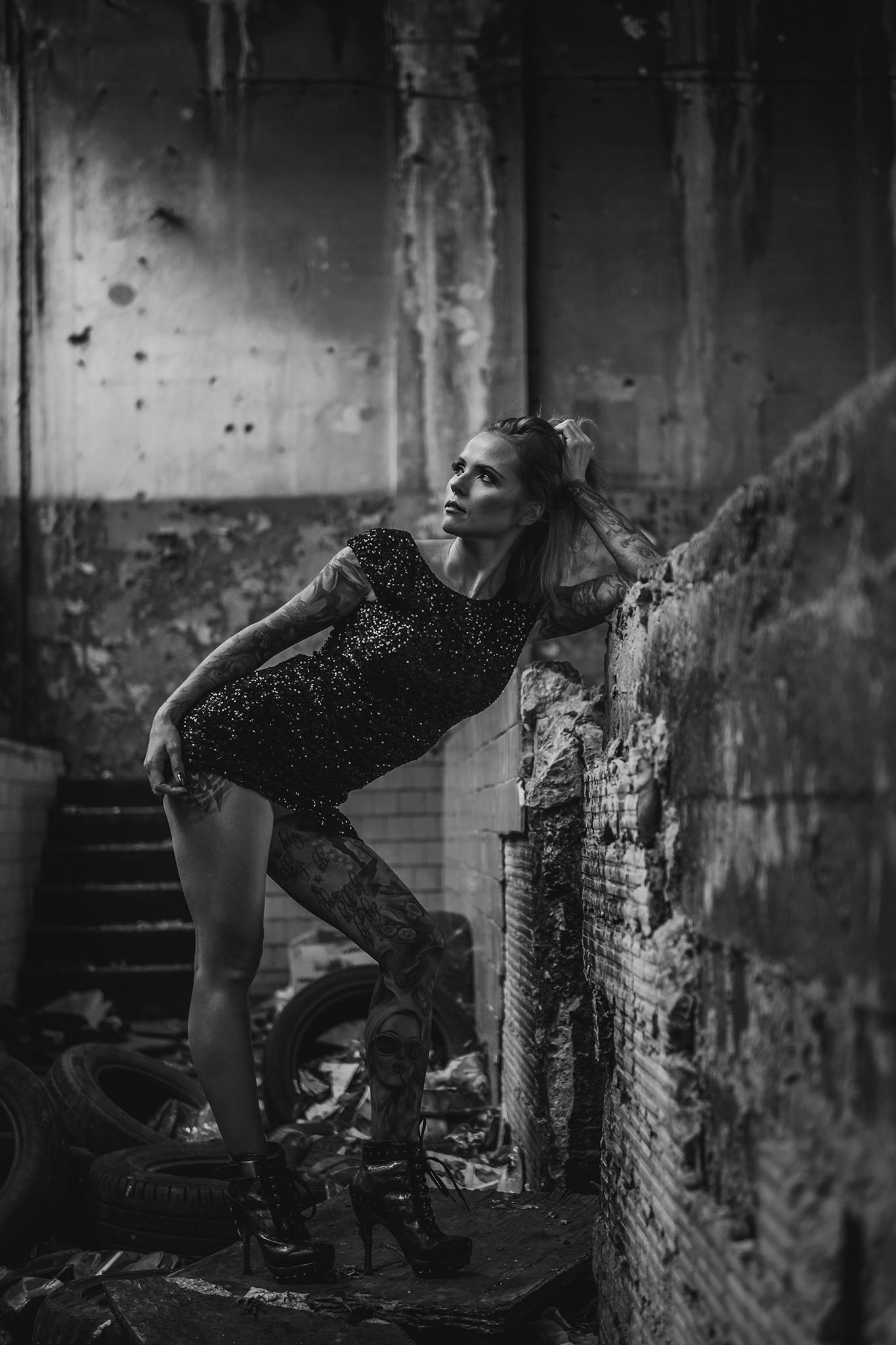 Czarno-biały portret kobiety w cekinowej sukience, opierającej się o zniszczoną ceglaną ścianę w opuszczonym wnętrzu, z widocznymi oponami i schodami w tle, eksponujący tatuaże na ciele modelki.