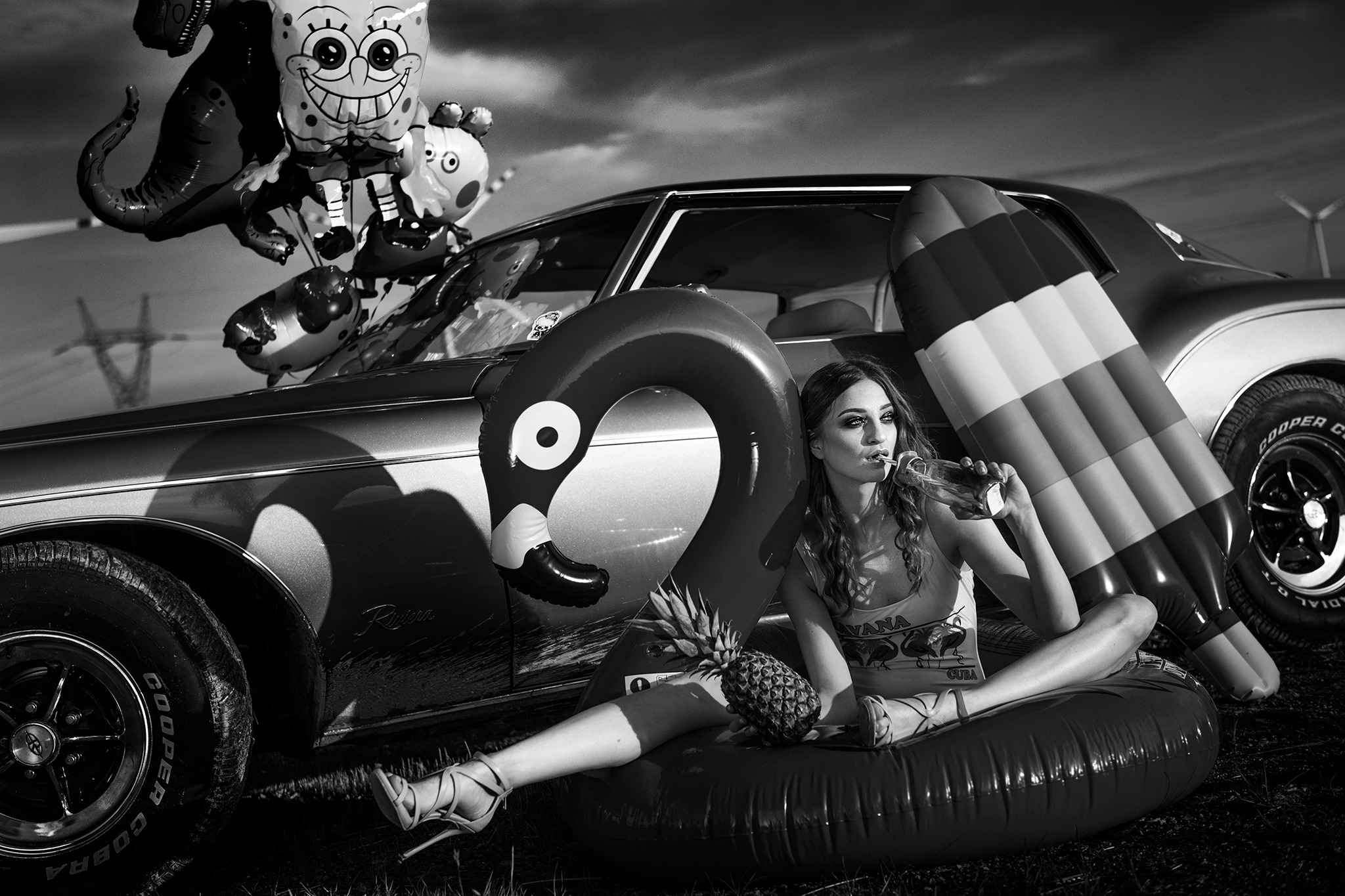 Czarno-białe zdjęcie kobiety siedzącej na dmuchanym flamingiem, pijącej z butelki, z ananasem na kolanach, obok klasycznego samochodu Buick Riviera z balonami i dmuchanym materacem, na tle...