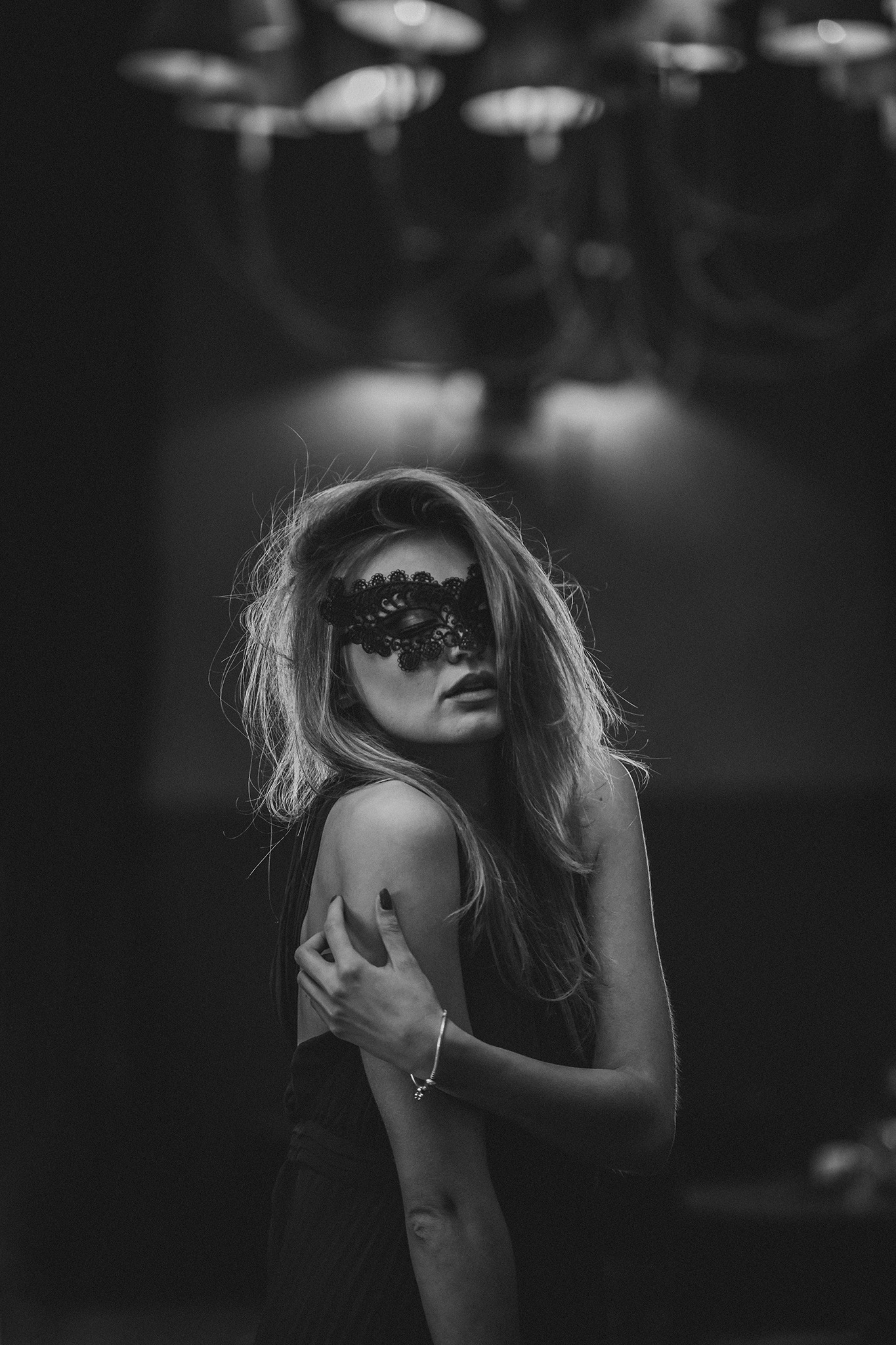 Czarno-biały portret kobiety w koronkowej masce na tle rozmytego oświetlenia, z ręką na ramieniu i srebrną bransoletką.
