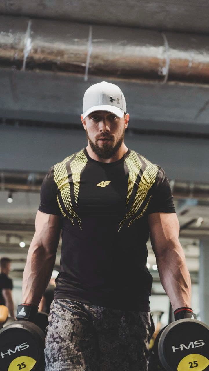 Mężczyzna z brodą w czarnej koszulce sportowej z żółtymi wstawkami i czapce z daszkiem, trzymający hantle w siłowni.