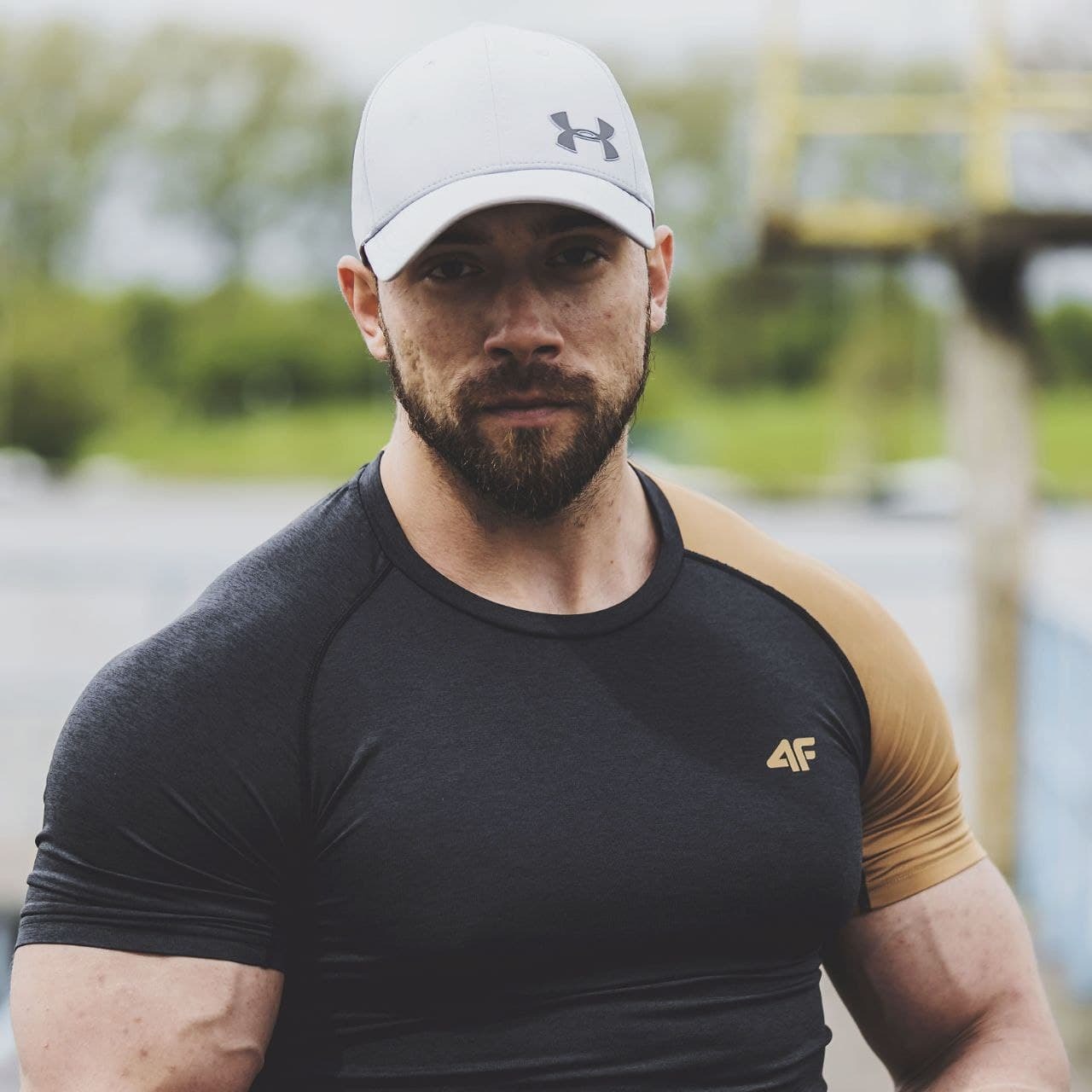 Mężczyzna w sportowej koszulce i czapce z daszkiem, z brodą, na tle boiska do koszykówki, patrzący wprost w obiektyw.