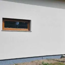 Fragment elewacji budynku z dwoma oknami o drewnianych ramach, widoczny kran na ścianie i fragment fundamentu z szarą izolacją.