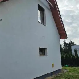 Boczny widok na świeżo pomalowaną białą elewację domu z dwoma małymi oknami, widoczne elementy systemu monitoringu, w tle nowoczesny budynek z ciemną blachą na dachu i zielone tuje.