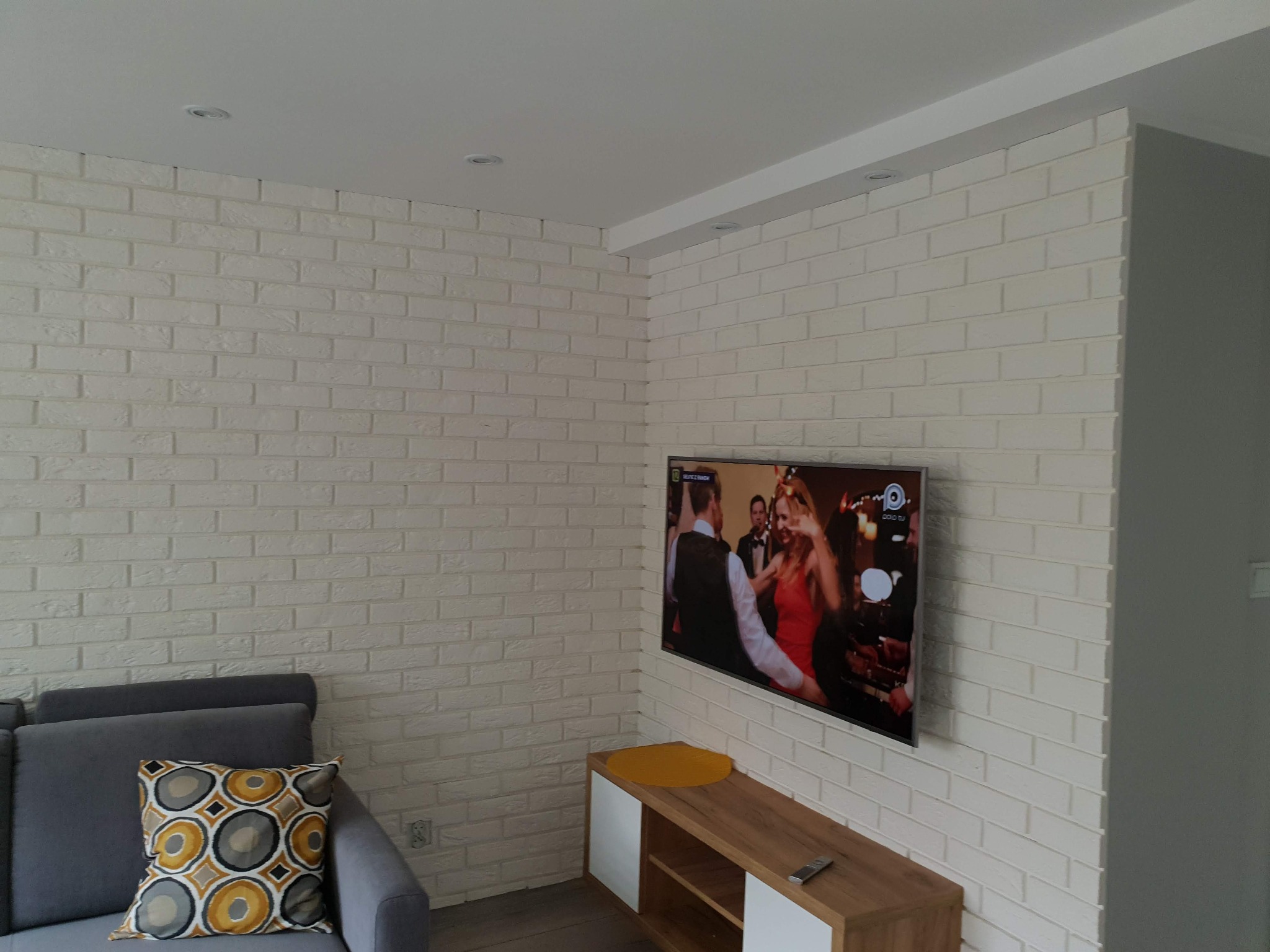 Ściana z białej cegły dekoracyjnej w salonie w Prudniku, podświetlona punktowymi lampami sufitowymi, z telewizorem i szarą sofą z wzorzystą poduszką.
