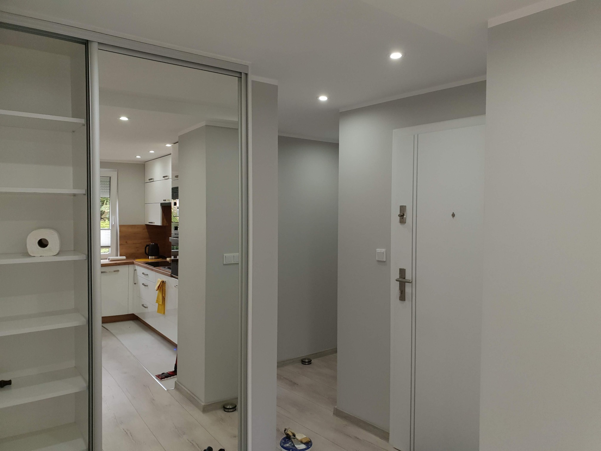 Wnętrze mieszkania po remoncie: widok z korytarza na kuchnię z białymi meblami i drewnianym blatem, białe ściany, drzwi wejściowe, szafa z lustrzanymi drzwiami przesuwnymi, na podłodze leżą...