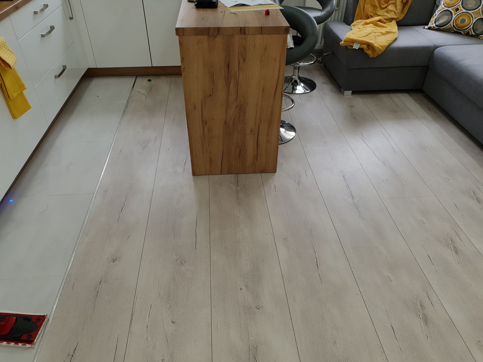 Jasne panele podłogowe z widoczną strukturą drewna, fragment kuchni z białymi szafkami, drewniany barek i szara kanapa z żółtym pledem w tle.