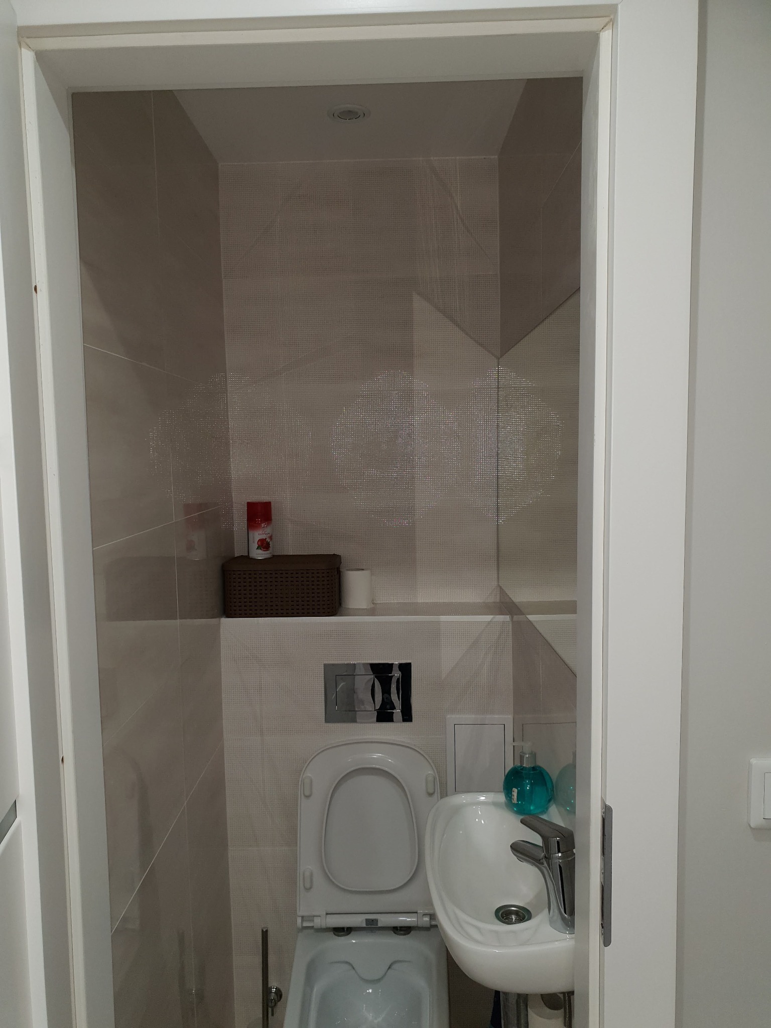 Wąska toaleta z umywalką, wykończona beżowymi płytkami z delikatnym wzorem, koszyk wiklinowy na półce, widoczna armatura i dozownik mydła w kolorze turkusowym.