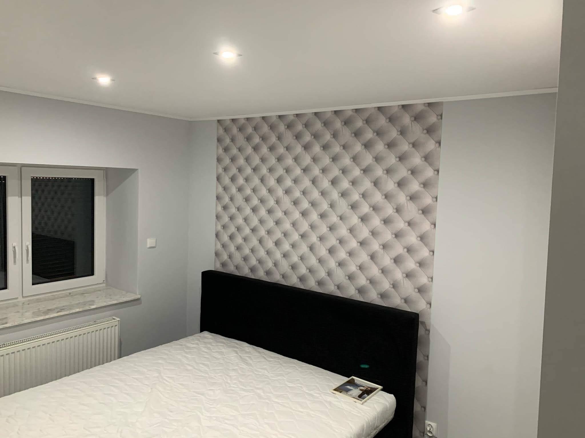 Sypialnia po remoncie: ściana z tapetą w stylu pikowanego materiału, czarne wezgłowie łóżka, biała pościel i grzejnik pod oknem.