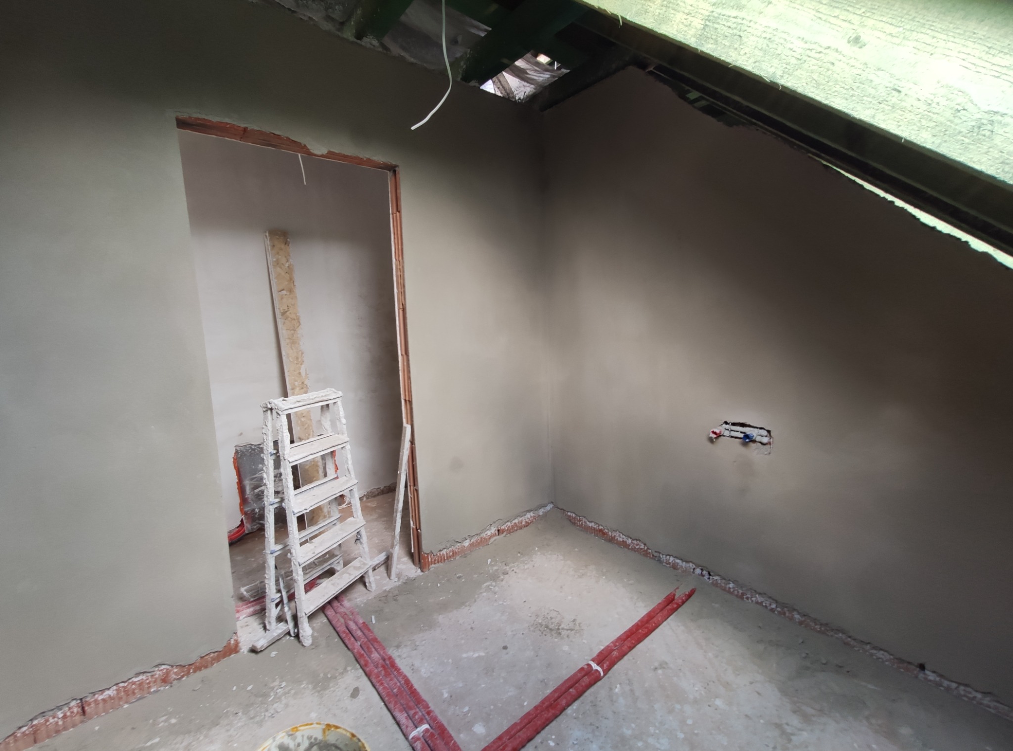 Pokój na poddaszu po nałożeniu tynków maszynowych, widoczna drabina malarska, czerwone rury grzewcze na podłodze, oraz otwór w suficie