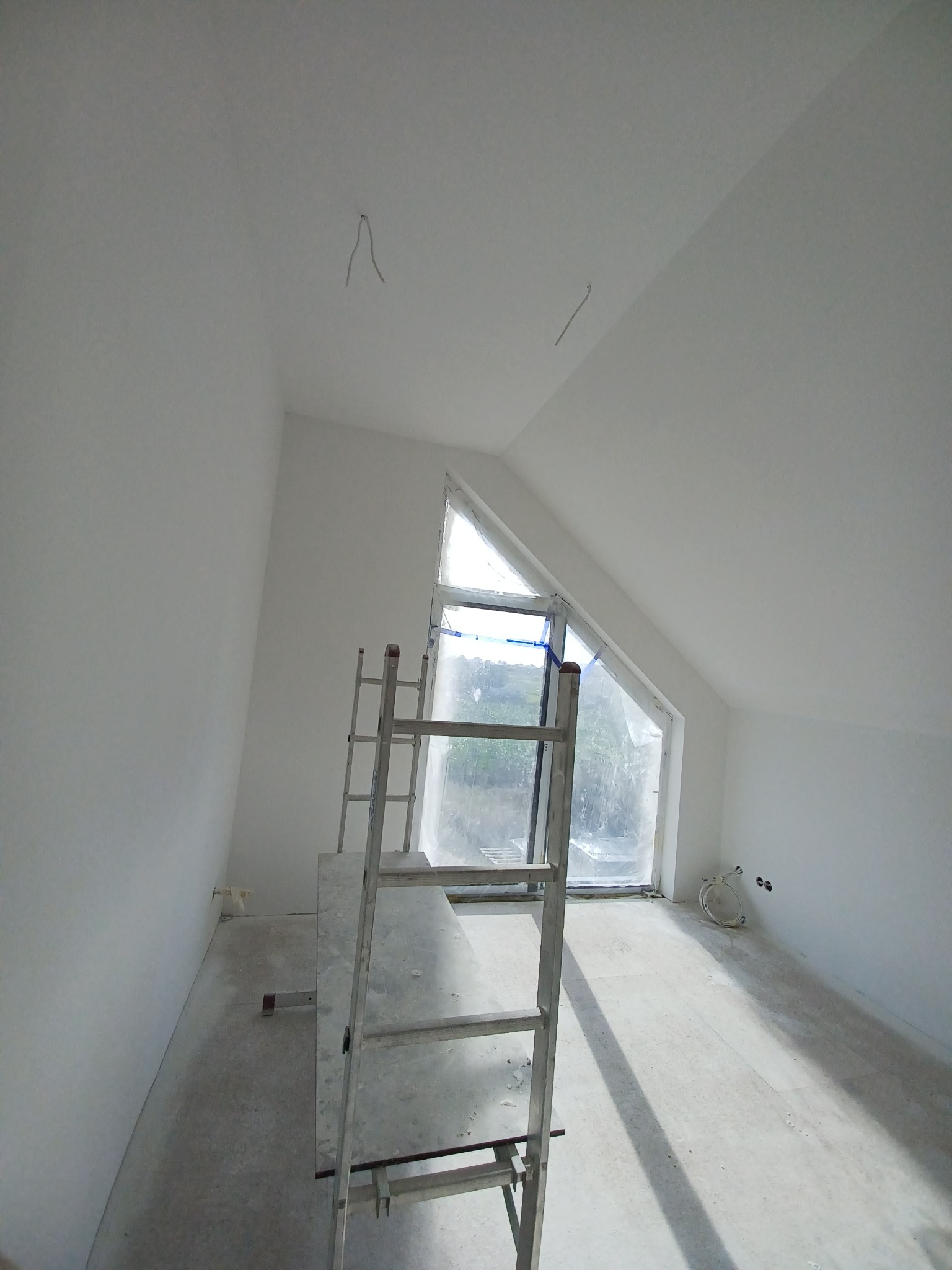 Poddasze po remoncie, świeżo pomalowane na biało, z zabezpieczonym oknem trójkątnym folią i rozstawionym rusztowaniem malarskim.