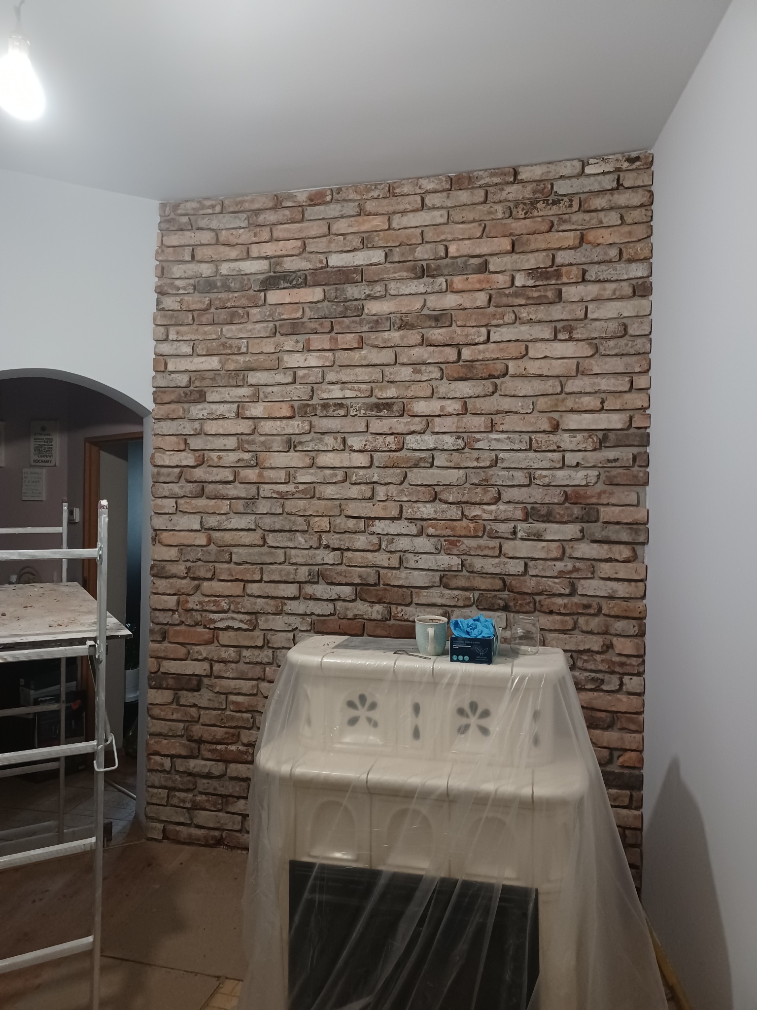 Ściana wykończona cegłą dekoracyjną w trakcie prac wykończeniowych, piec kaflowy zabezpieczony folią, widoczne narzędzia i drabina, w tle fragment pomalowanej ściany.