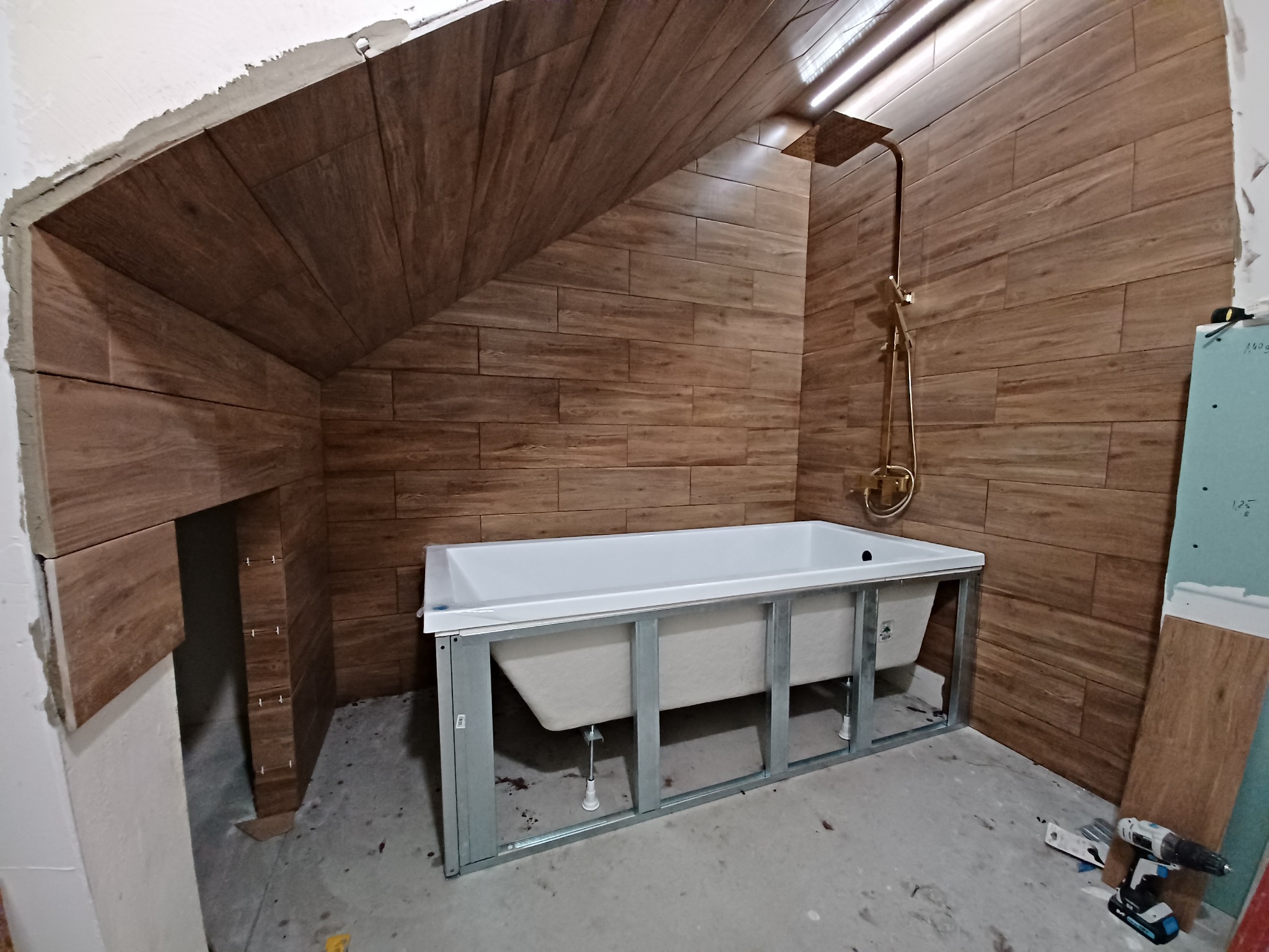 Montaż wanny na metalowym stelażu w łazience z drewnopodobnymi płytkami ściennymi, widoczny prysznic w kolorze złotym i narzędzia na podłodze.