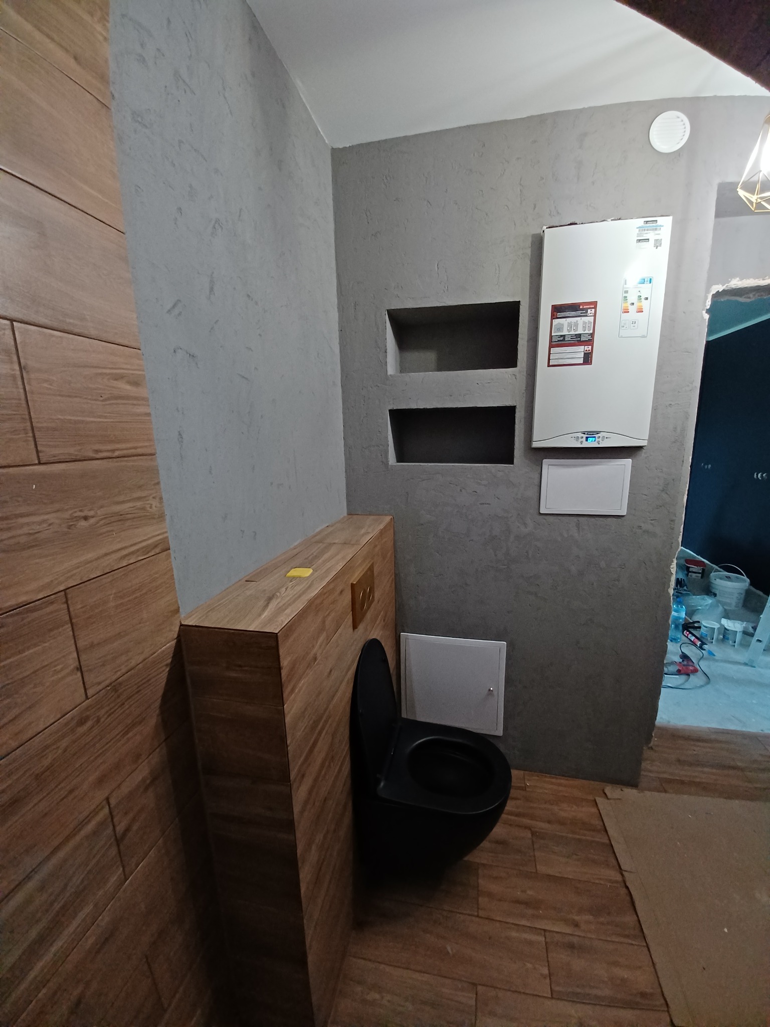 Wykończona toaleta z szarą ścianą o fakturze, drewnopodobnymi płytkami na ścianie i podłodze, oraz zabudowaną toaletą z czarną miską. Na ścianie widoczny piec gazowy i wnęki.