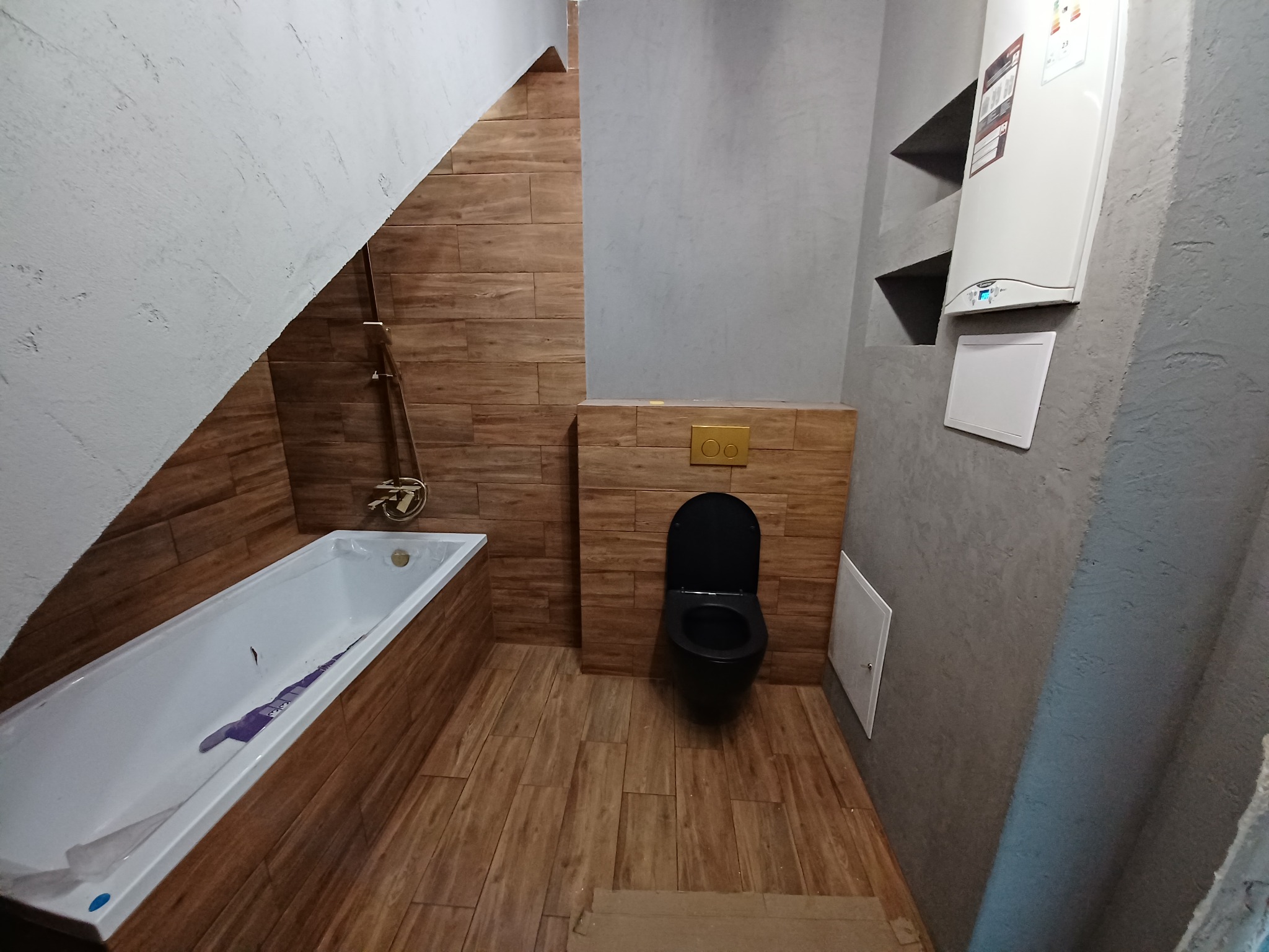 Wykończona łazienka na poddaszu z drewnopodobnymi płytkami, czarną miską ustępową z złotym przyciskiem spłukiwania, białą wanną z folią ochronną i szarymi ścianami, na których wisi biały piec gazowy.