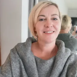 Uśmiechnięta blond kobieta w szarym swetrze robi selfie w lustrze, trzymając telefon w dłoni.