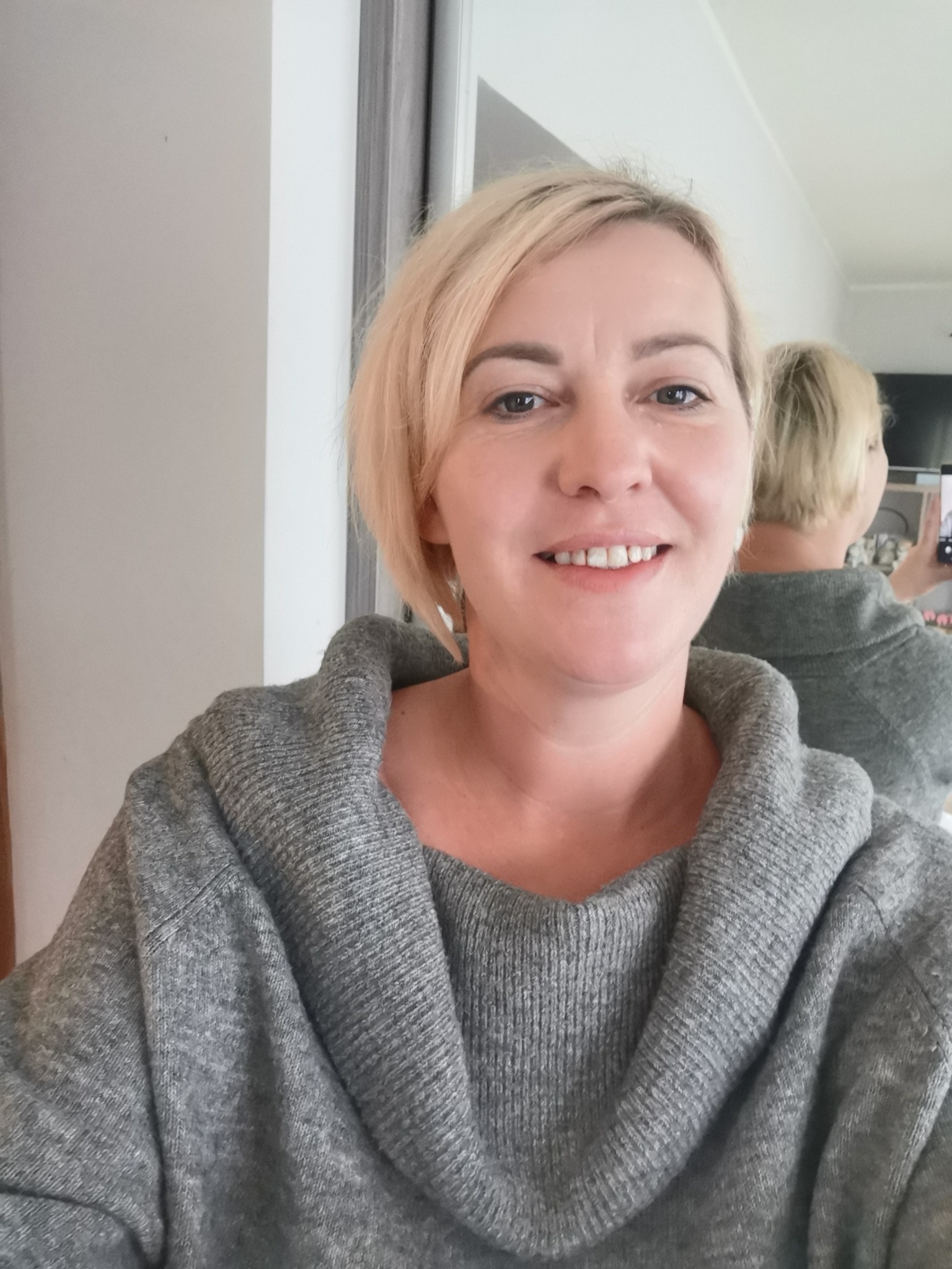 Uśmiechnięta blond kobieta w szarym swetrze robi selfie w lustrze, trzymając telefon w dłoni.